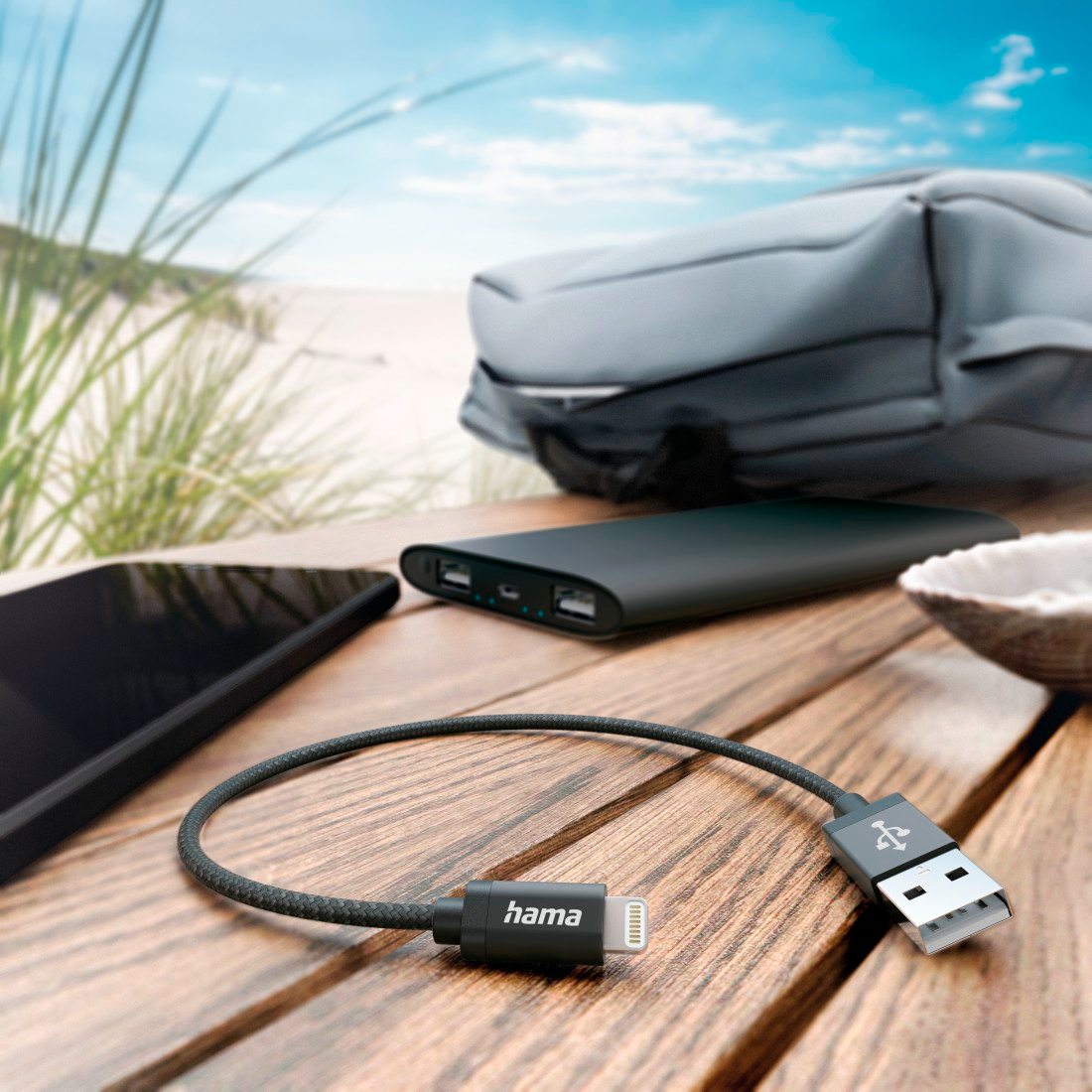 USB kábel telefonnal, power bank-kel és egy tengerparttal a háttérben.