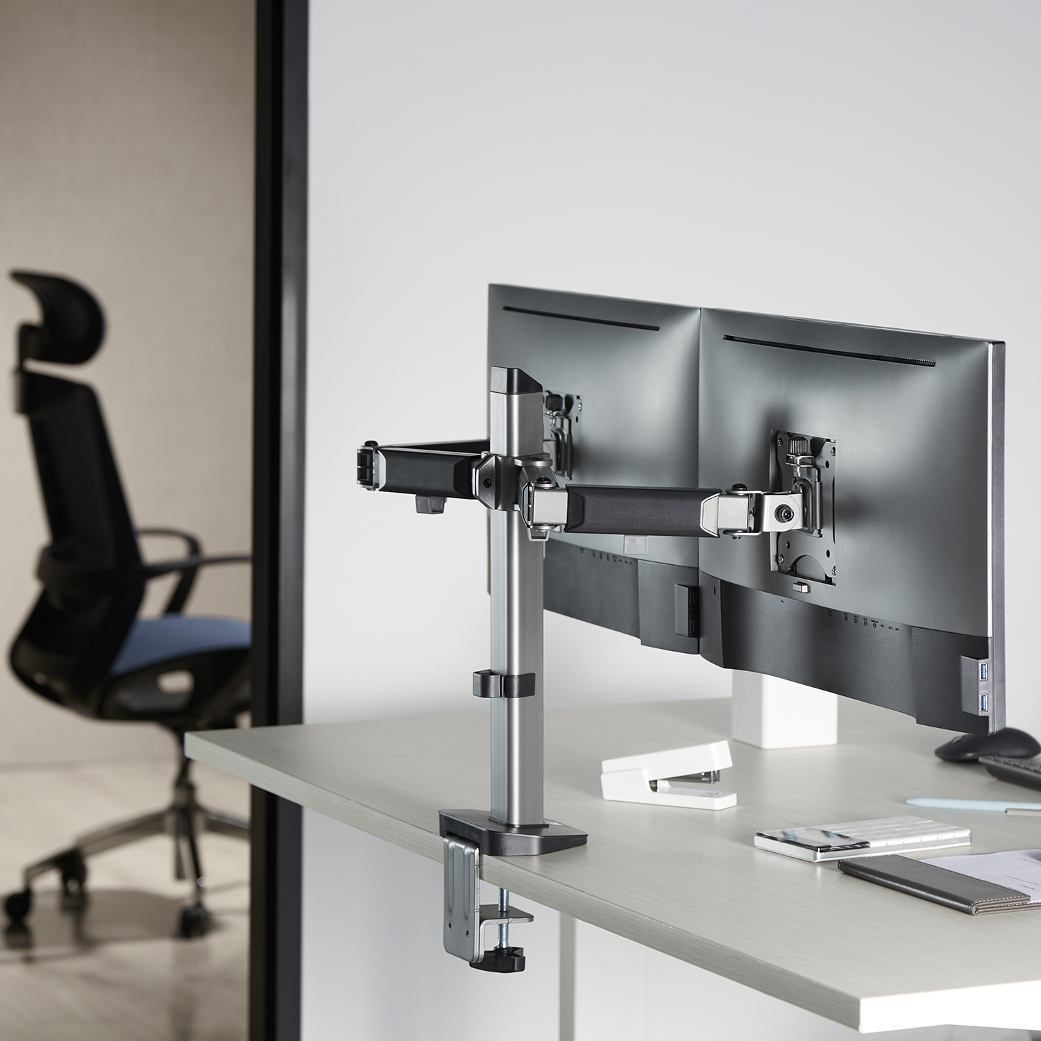 Dual monitor stand on a desk. Silver and black, with a chair and window in the background.