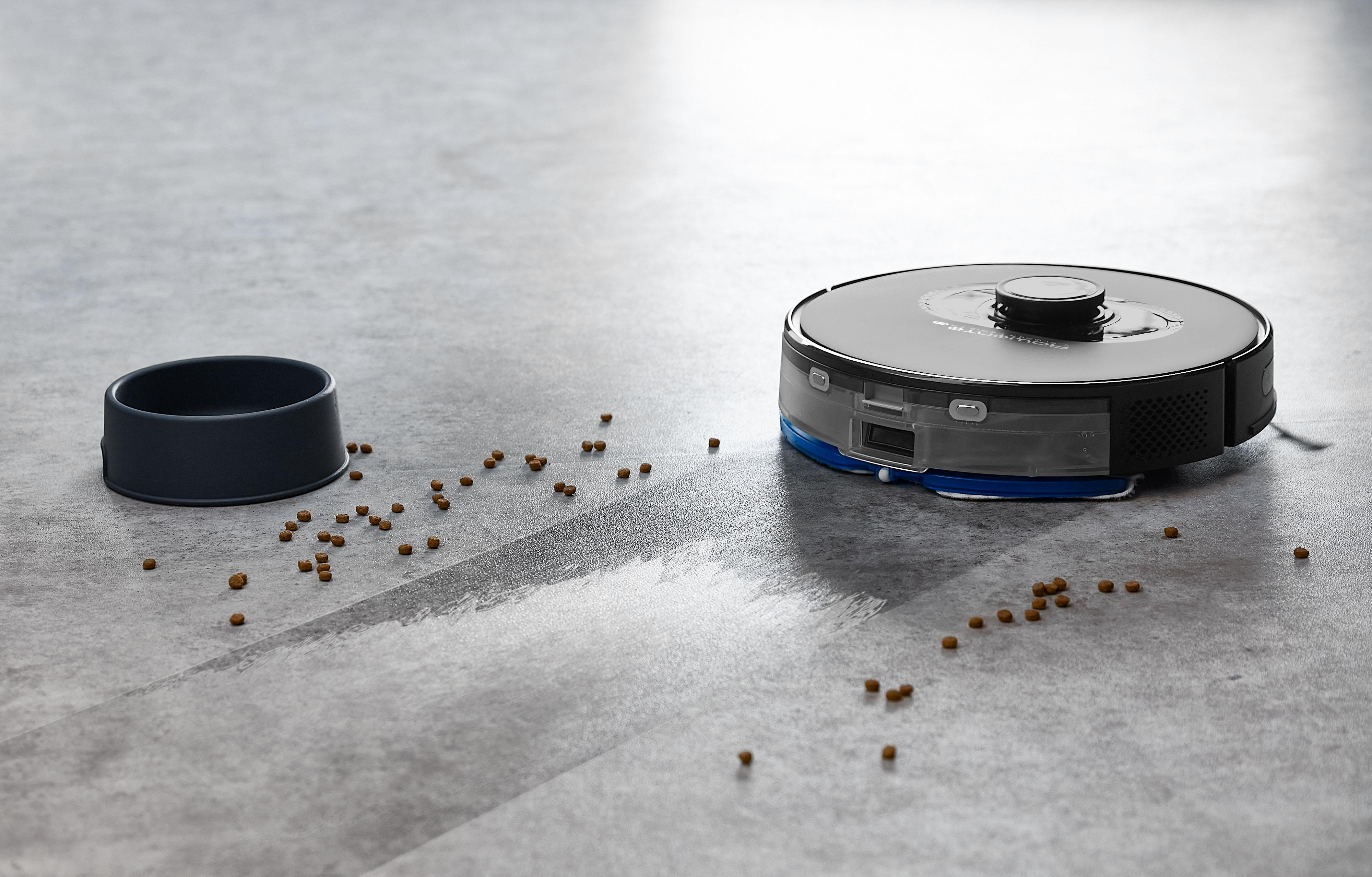 Ein schwarzer Roboterstaubsauger mit auf dem Boden verstreutem Essen und einer schwarzen Schüssel.