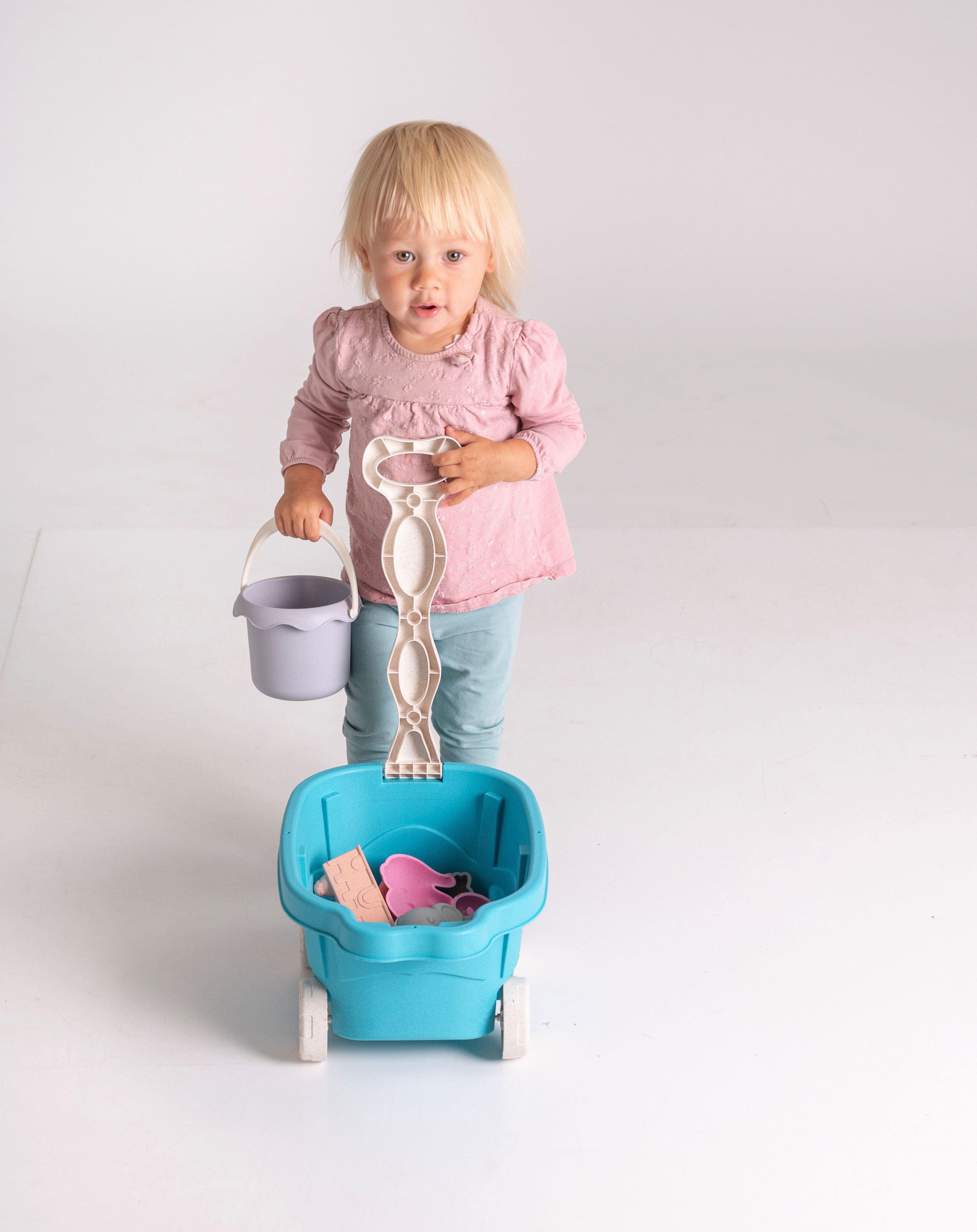 Ein junges Mädchen hält einen Eimer und zieht einen blauen Wagen mit Spielzeug.