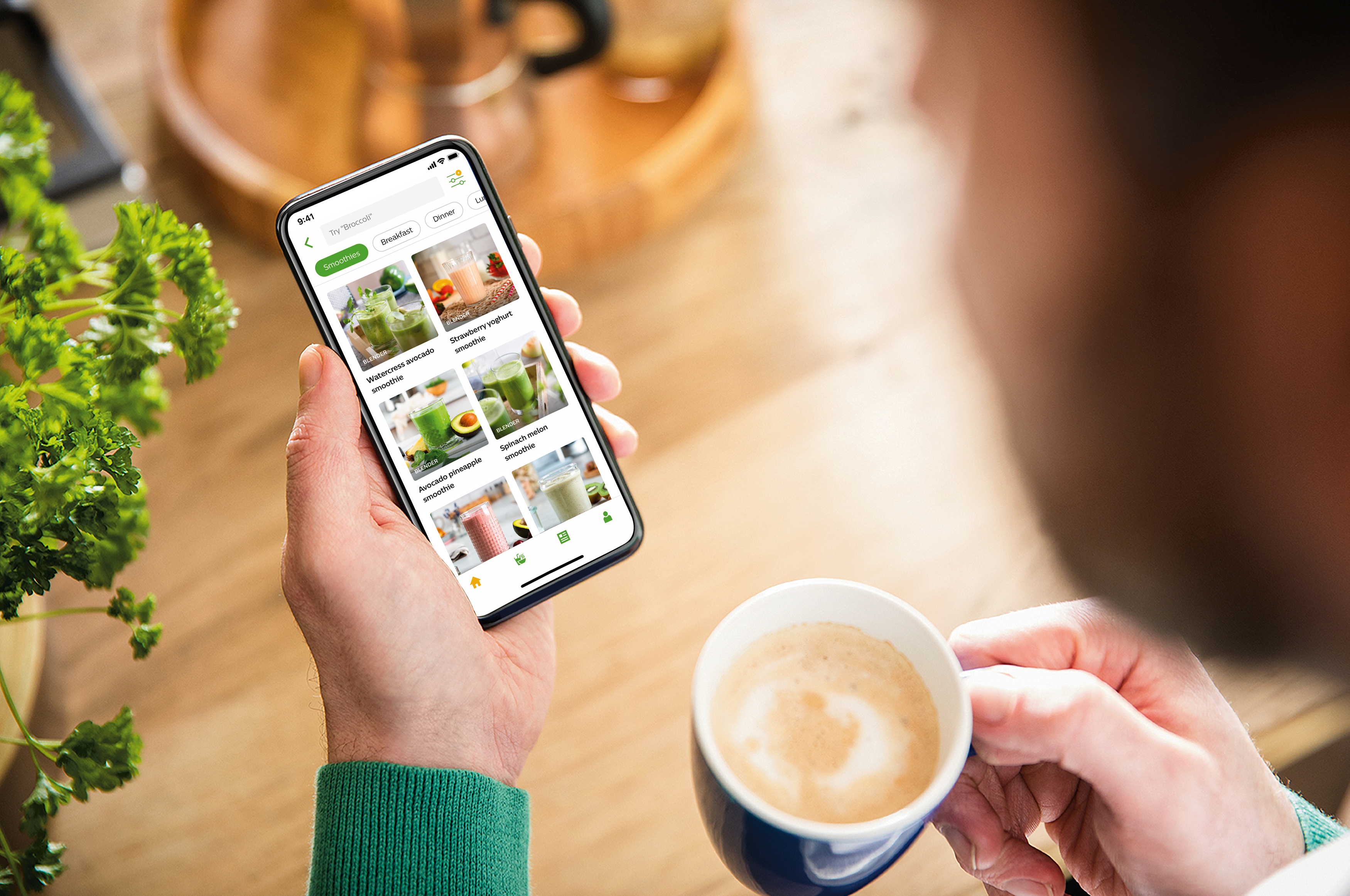 Una persona sosteniendo un teléfono inteligente que muestra una aplicación de recetas de batidos, con una taza de café.