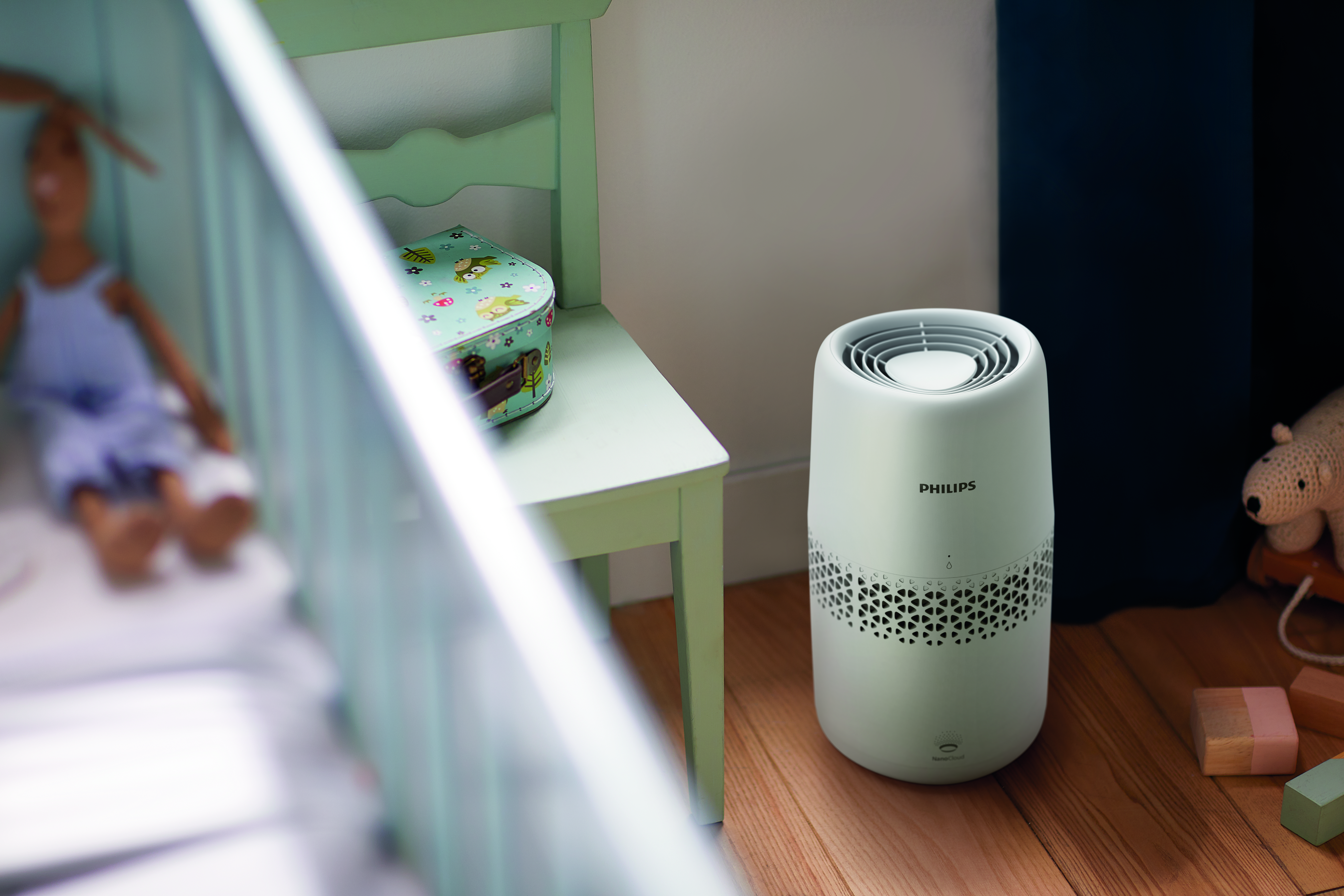 Un purificador de aire Philips blanco está junto a una silla con una caja decorada. Una cuna y un juguete están en el fondo borroso.