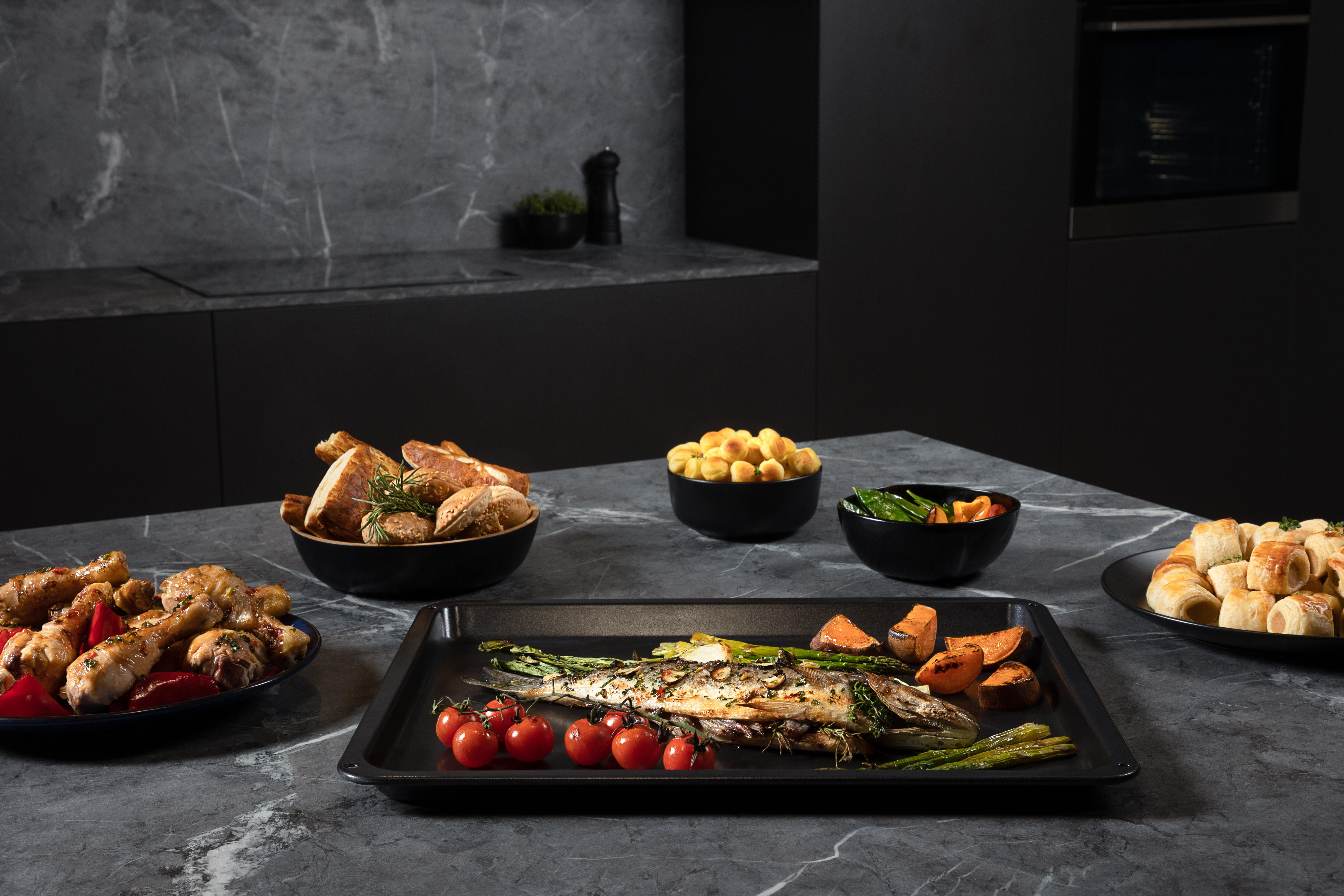 Küchenszene mit Essen auf einem Tisch, darunter Backwaren, Gemüse und gegrillter Fisch.