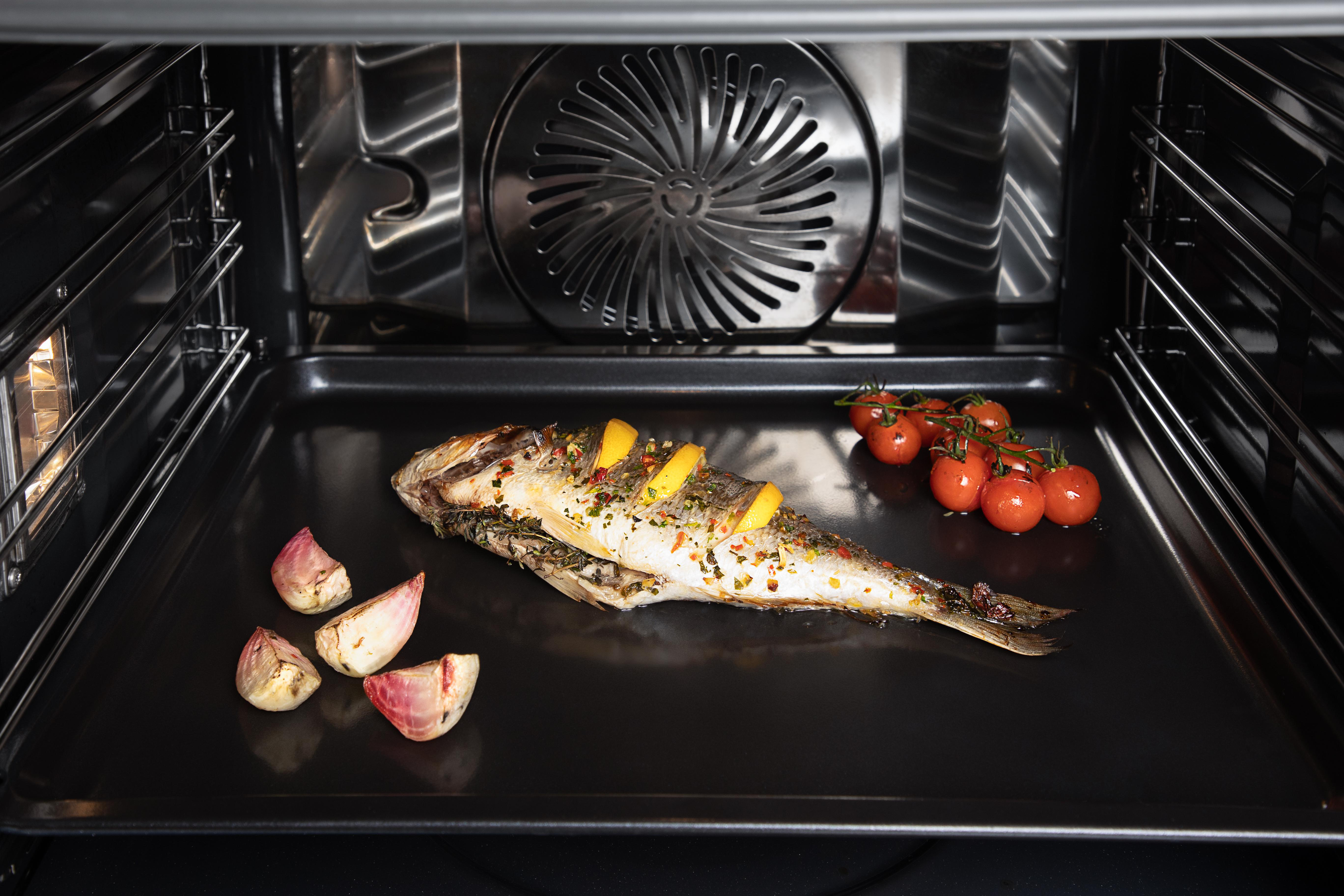 Gebackener Fisch mit Zitronenscheiben und Tomaten auf schwarzem Backblech.