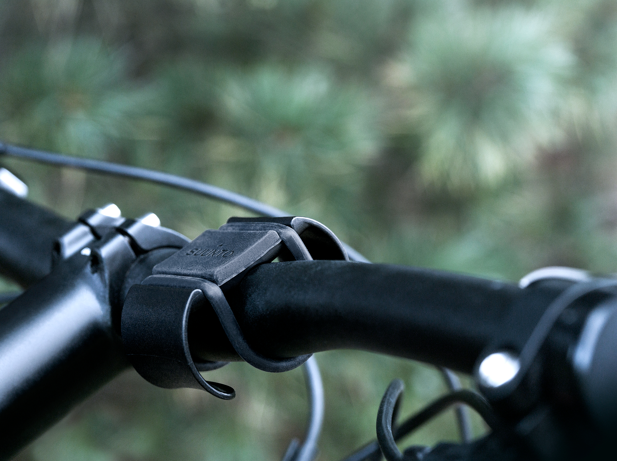 Nahaufnahme eines schwarzen Fahrradlenkers mit einem schwarzen Gerät. Grüne Bäume im Hintergrund.