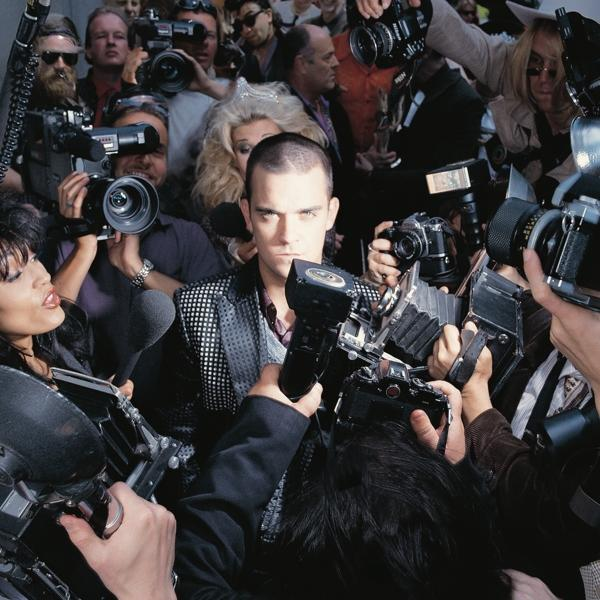 Robbie Williams omringd door paparazzi met camera's.