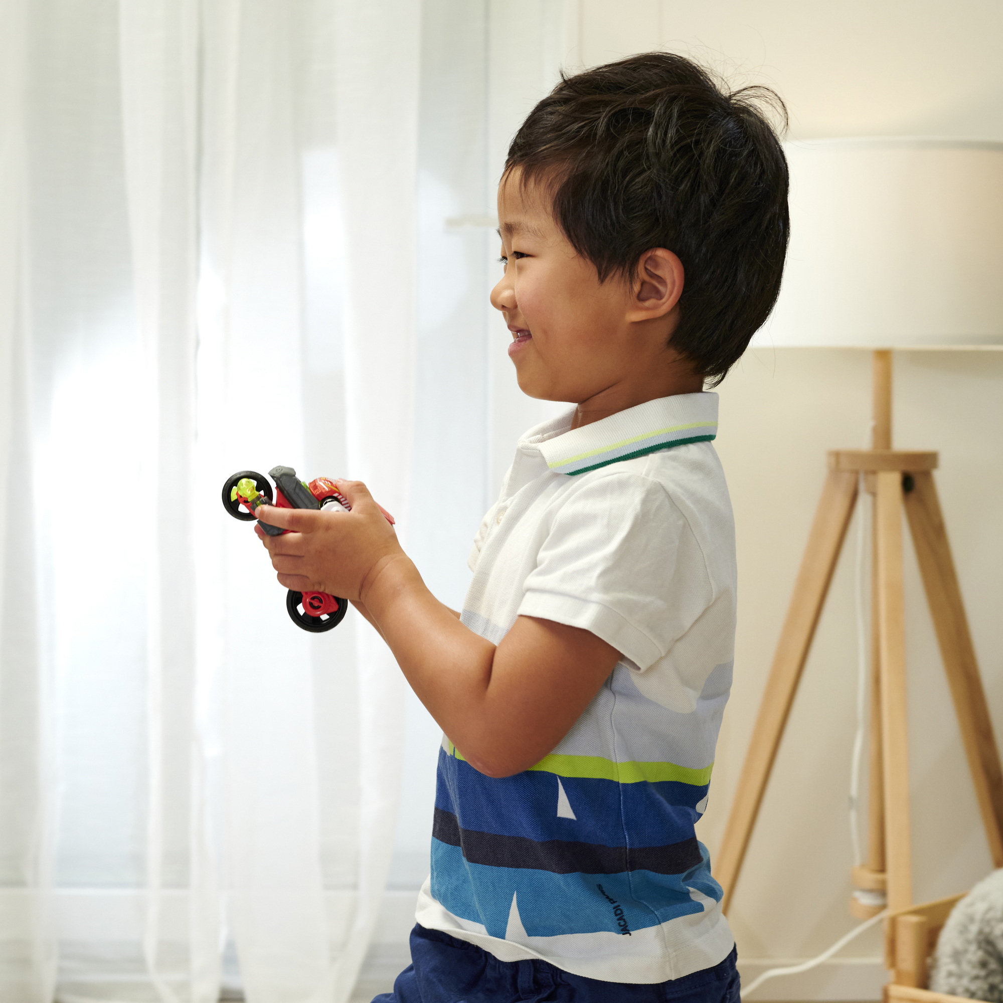 Ein junger Junge hält ein Spielzeugauto und lächelt. Der Hintergrund umfasst ein Fenster und eine Lampe.