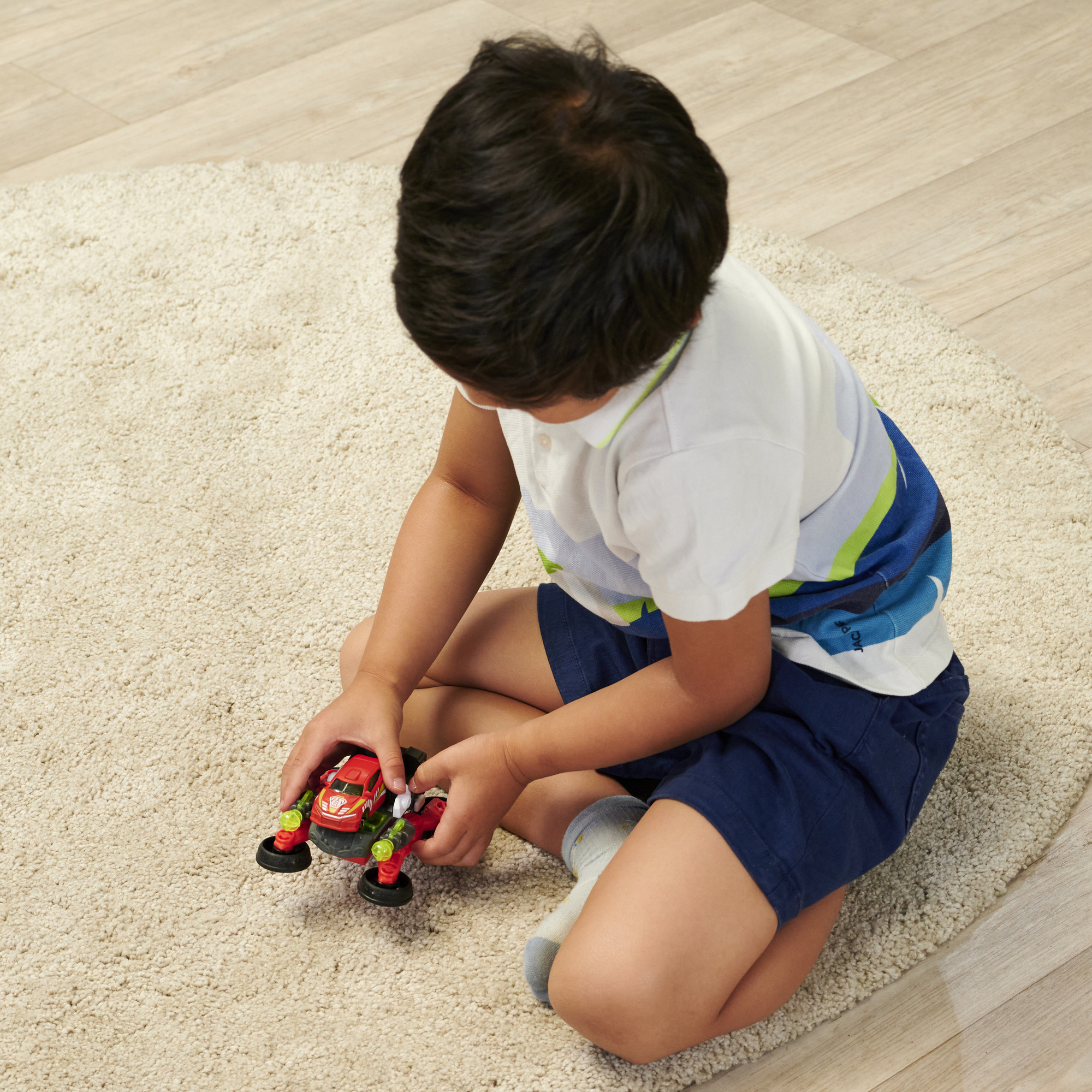 Kind spielt mit rotem und grünem Spielzeugauto auf cremefarbenem Teppich. Kind sitzt.
