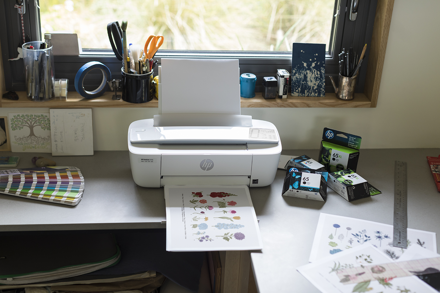White printer with paper and ink cartridges on a gray desk with craft supplies.