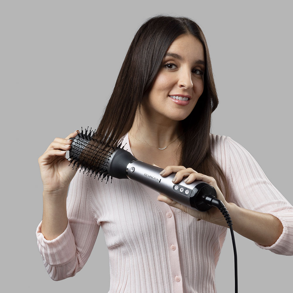Nő a haját formázza egy Remington levegős hajformázóval, a kamerába mosolyog.
