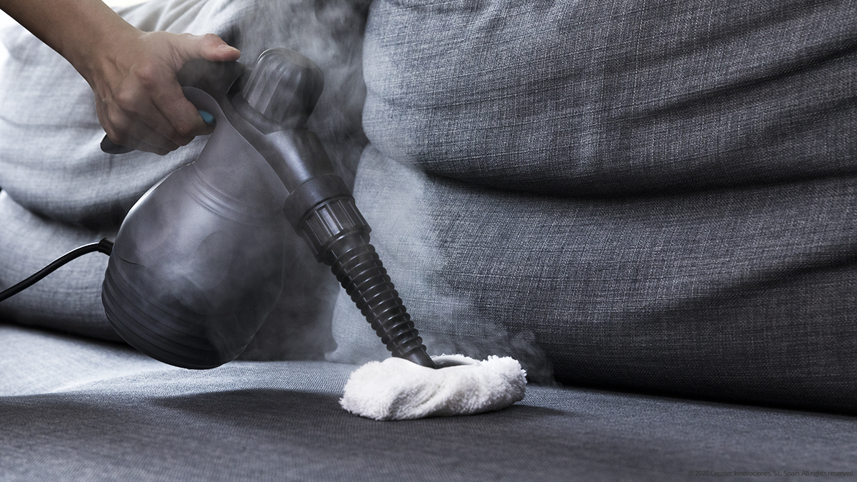 Una mano usando un limpiador a vapor en un sofá gris. Vapor visible.
