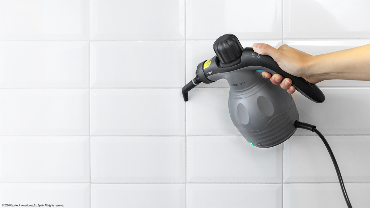 La mano de una persona limpiando una pared de azulejos blancos con un limpiador a vapor gris.