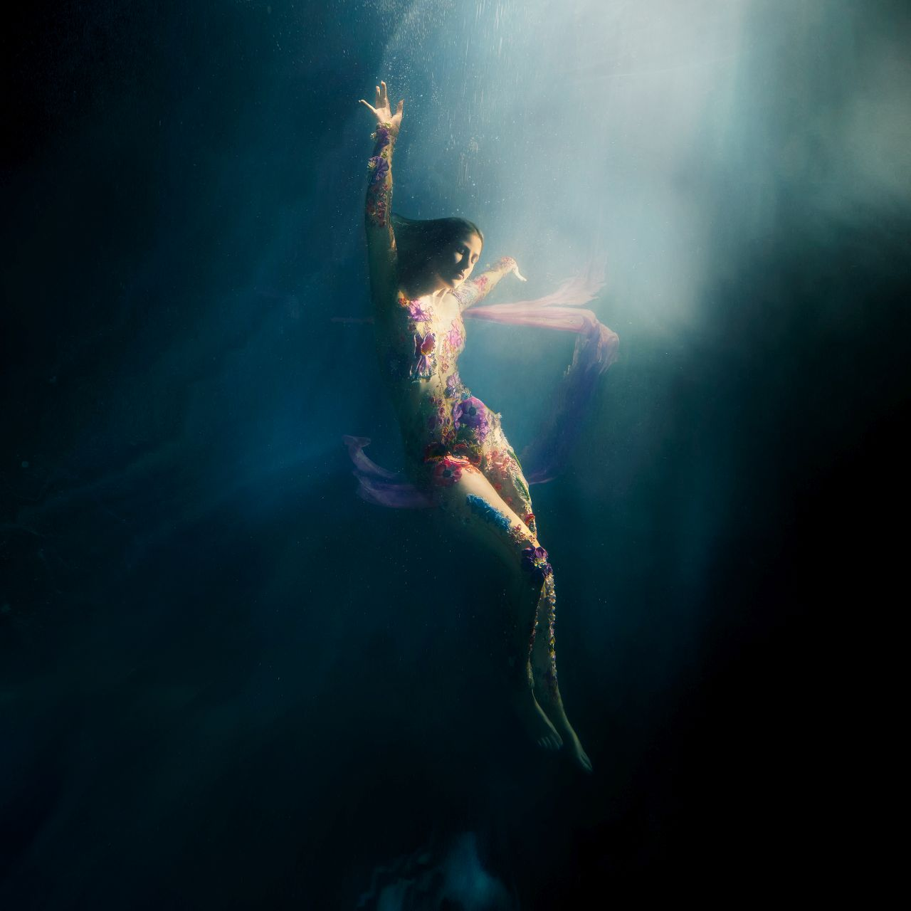 Eine Frau in einem Blumendress unter Wasser, Arme zum Licht erhoben. Der Hintergrund ist dunkel.