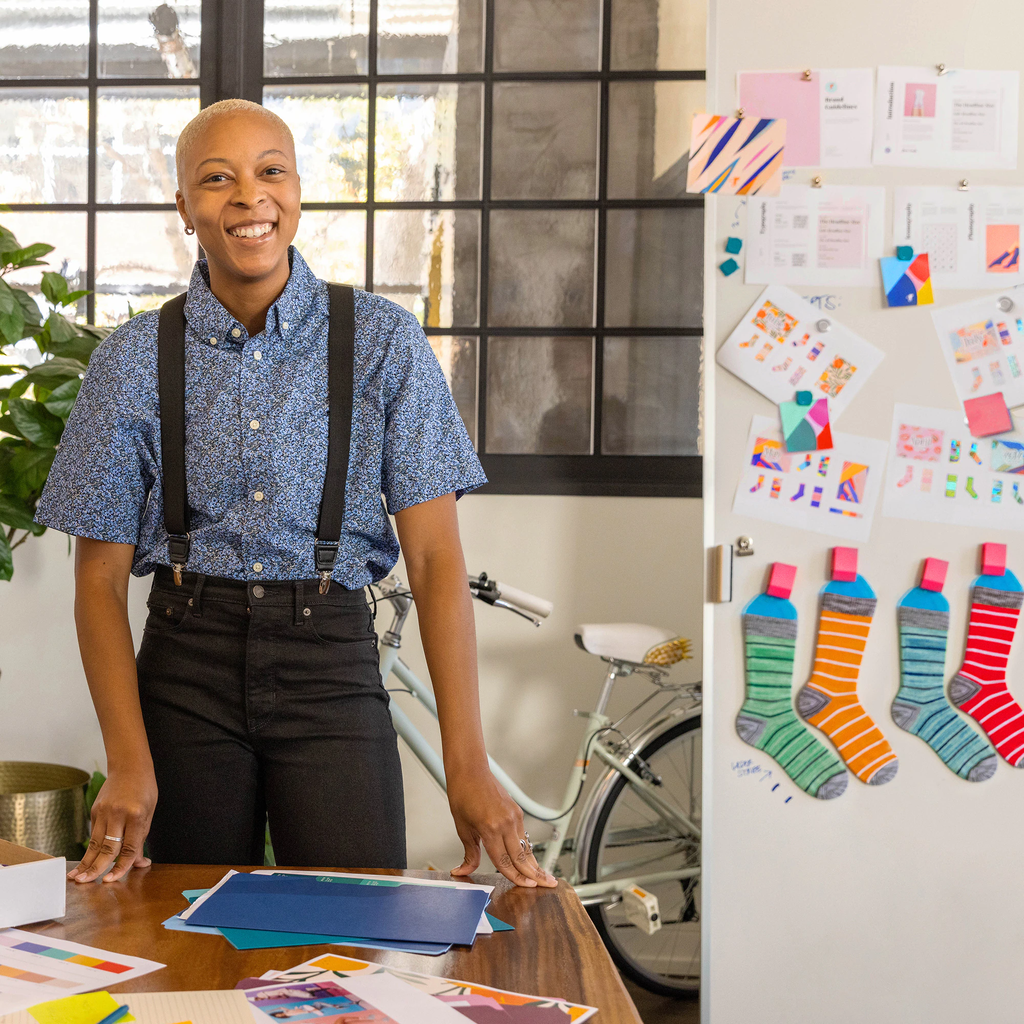 Femme en chemise bleue et pantalon noir debout dans un bureau, souriante.
