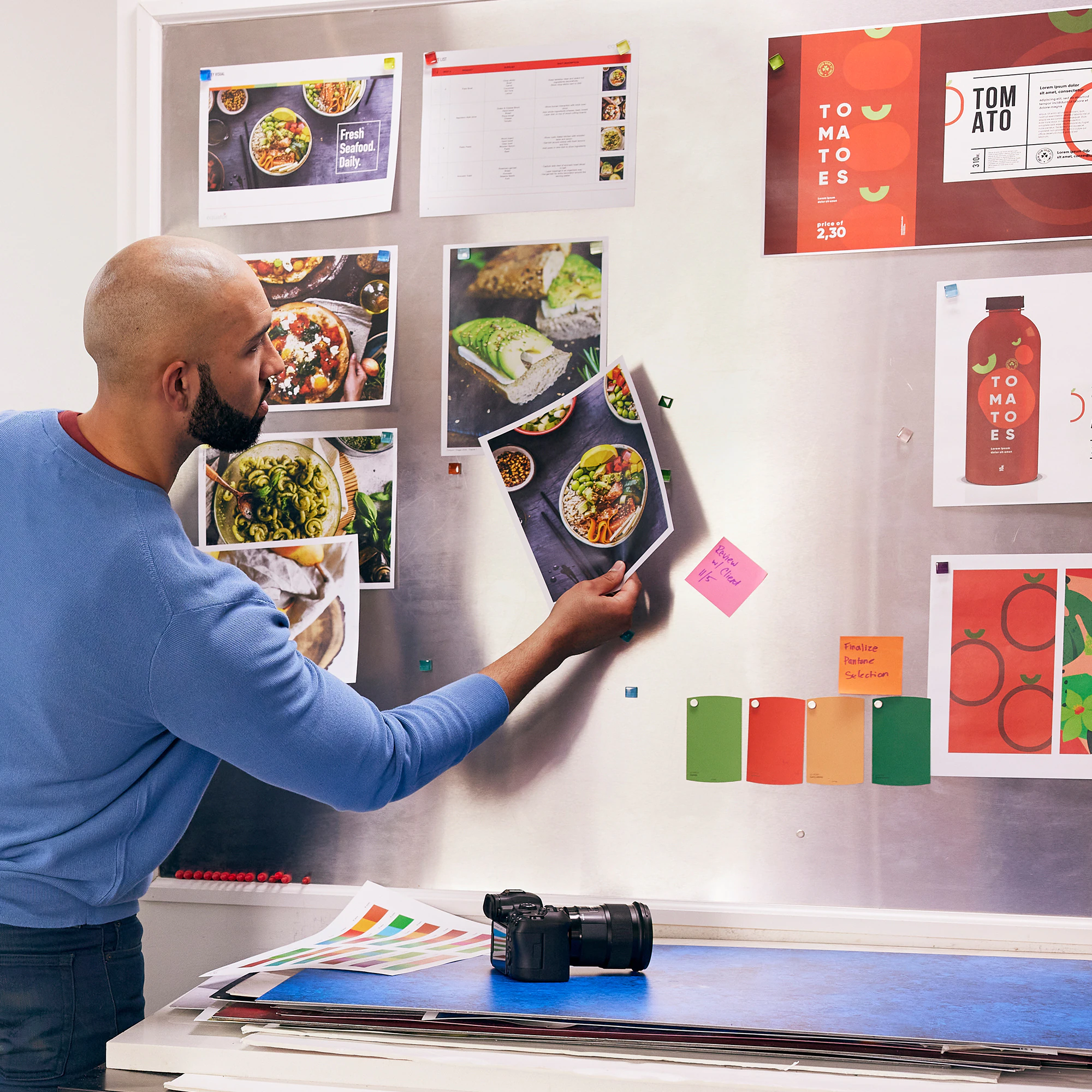 Un homme regarde des images de nourriture sur un tableau métallique. Autres éléments:échantillons de couleurs, appareil photo et papiers.