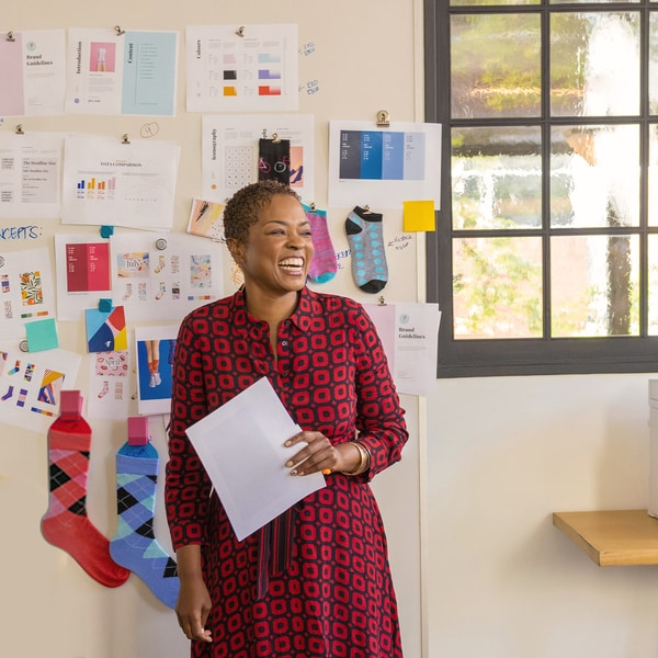 Femme souriante en robe rouge à motifs devant un mur couvert de papiers et de chaussettes.