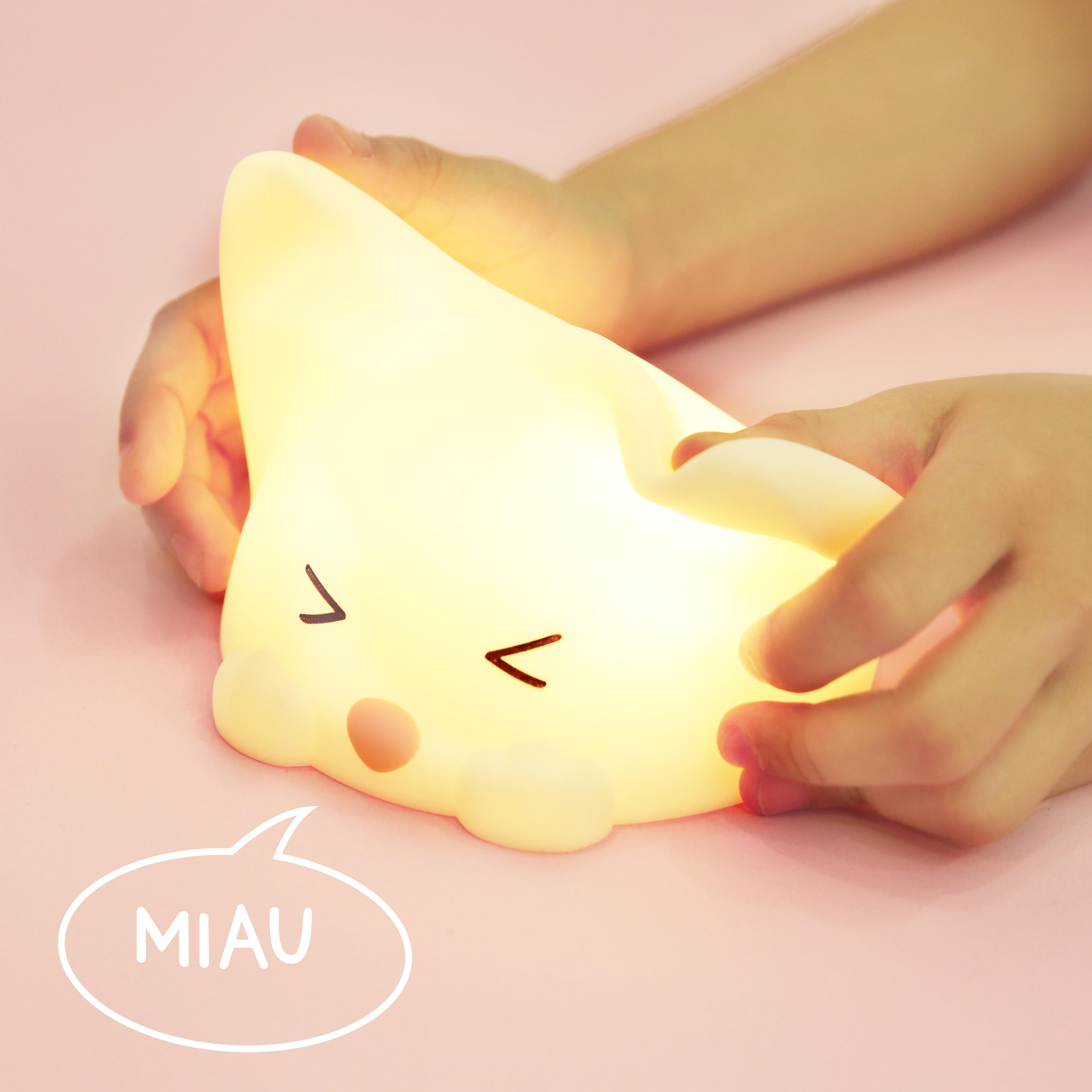 Eine katzenförmige Nachtlampe, die von Händen auf einer rosa Oberfläche gehalten wird, mit einer Sprechblase 'MIAU'.