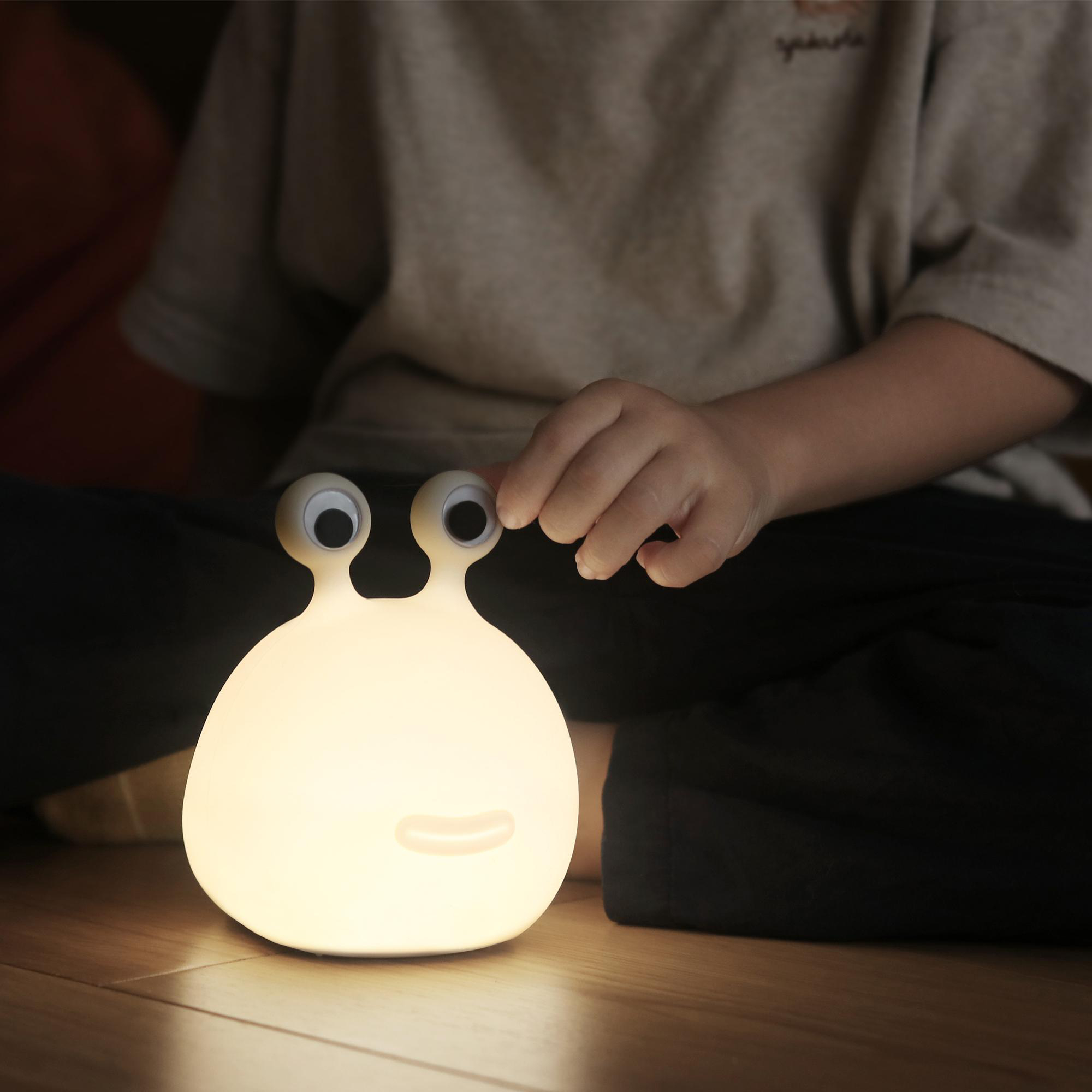 Glühende weiße Lampe mit großen Augen, die von der Hand eines Kindes berührt wird. Die Lampe steht auf einem Holzboden.