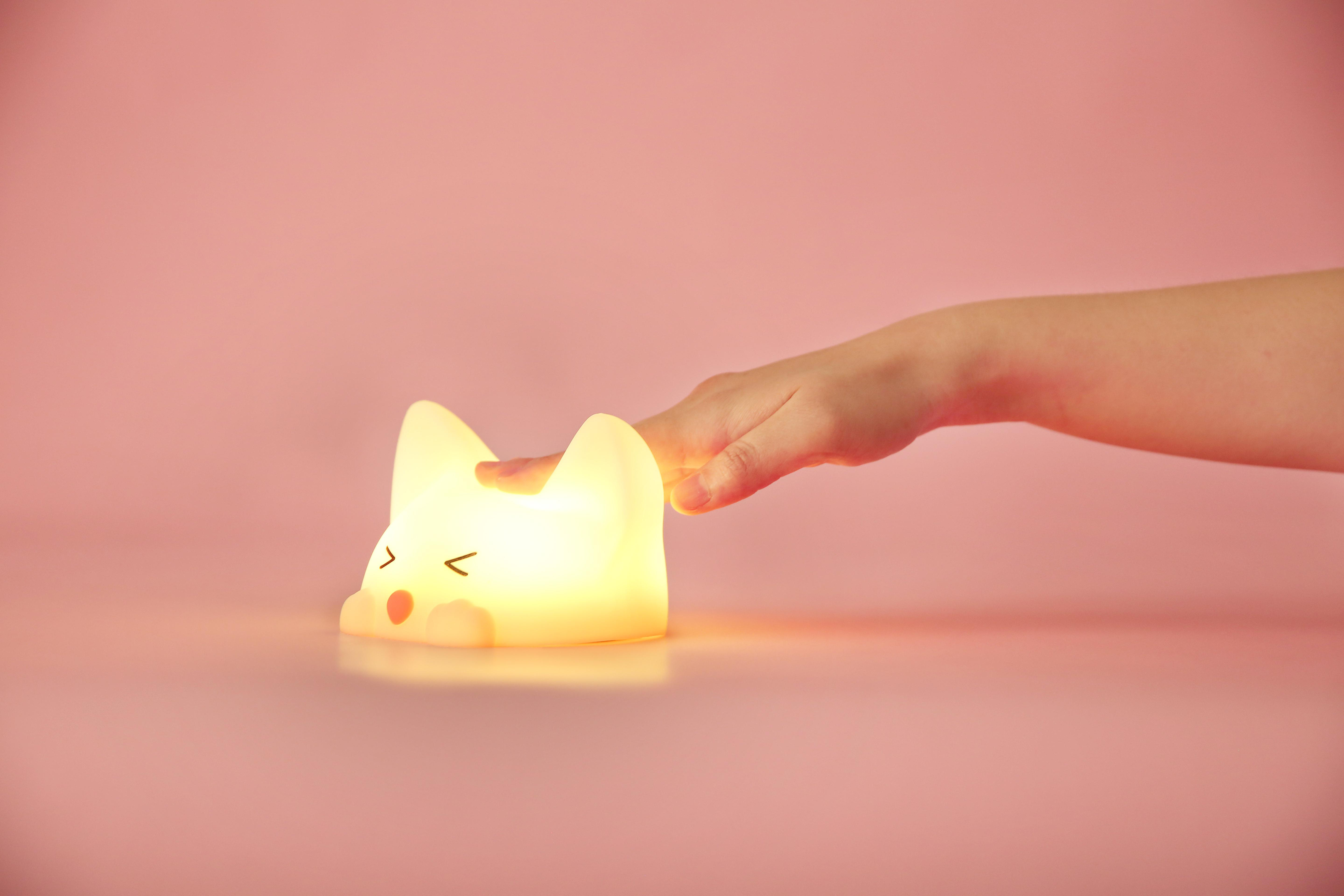 Eine Hand berührt eine leuchtende, weiße katzenförmige Lampe auf rosa Untergrund.