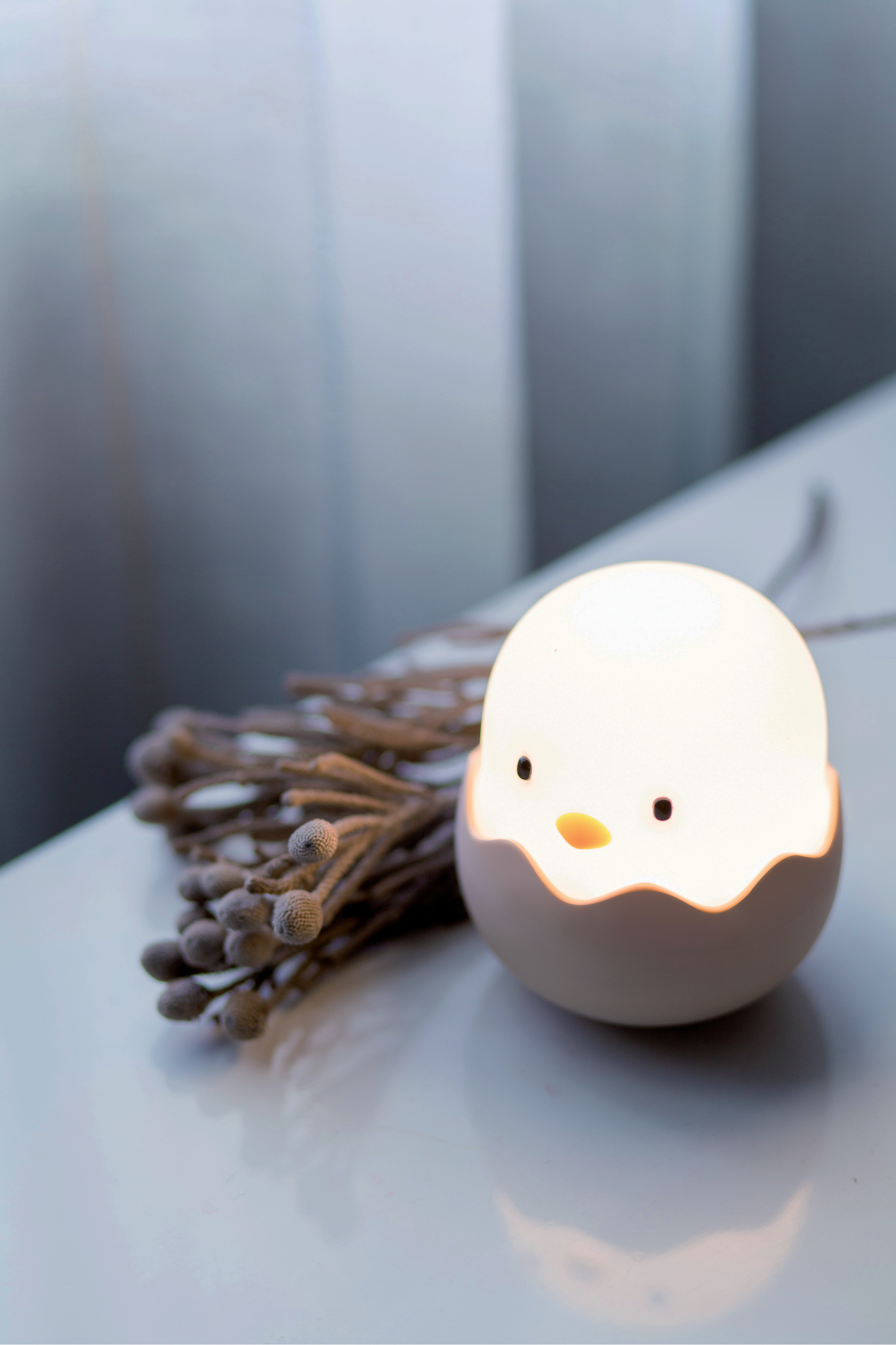 Eiförmige Lampe mit Küken-Gesicht, auf weißer Oberfläche mit Zweig. Weiche Beleuchtung.
