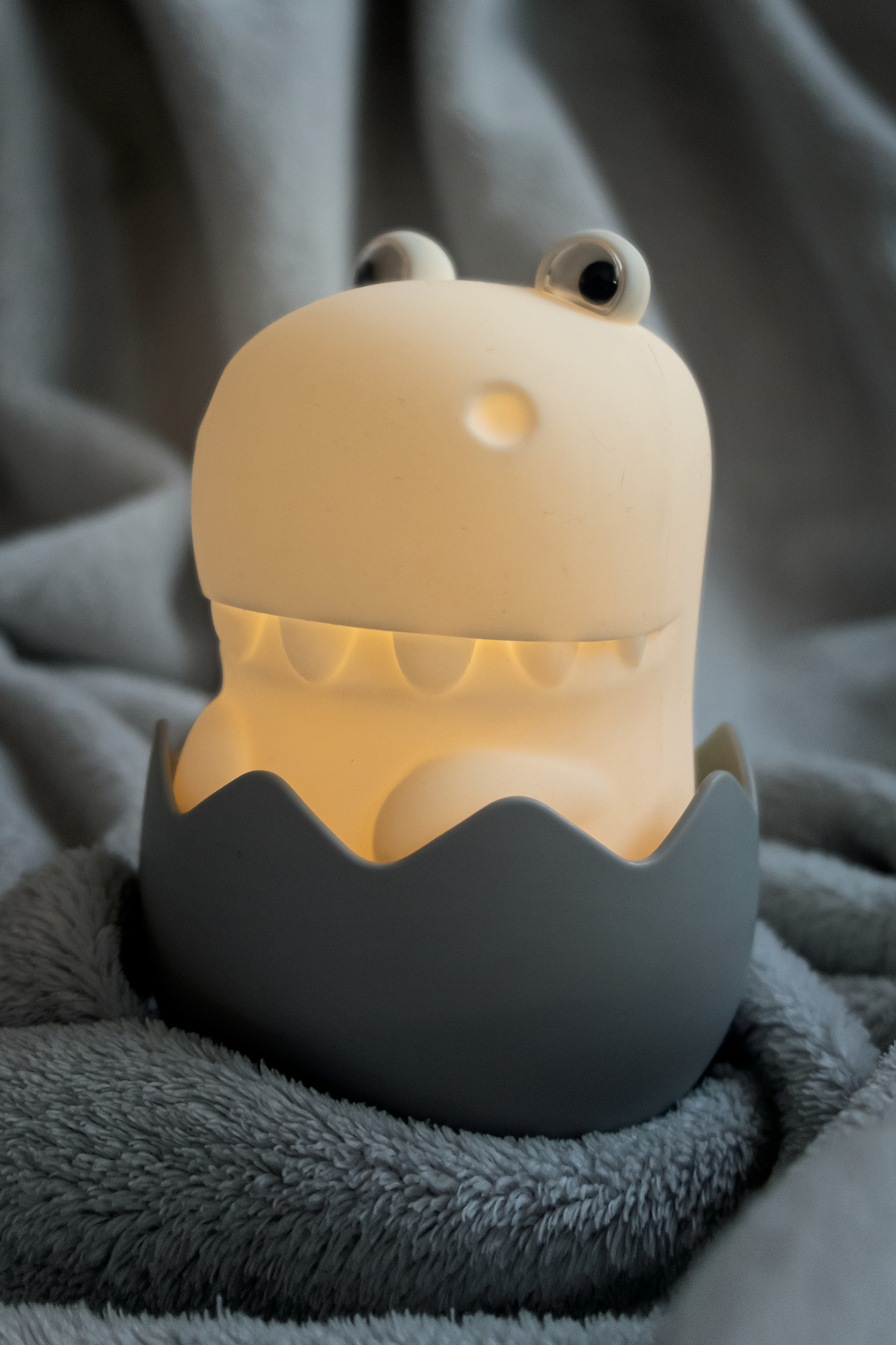 Ein Dinosaurier-Nachtlicht in einem grauen Ei, auf einer grauen, flauschigen Decke.