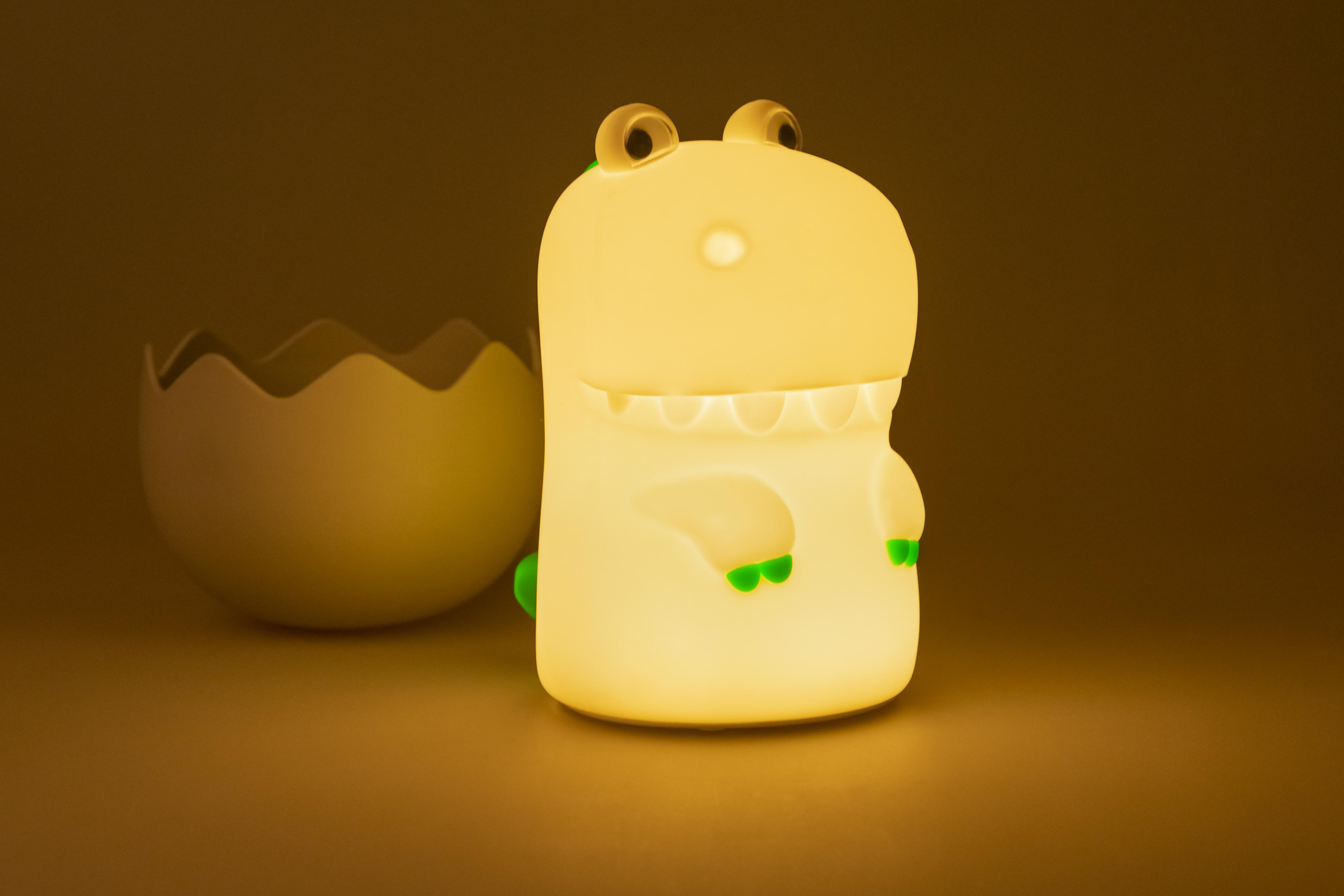 Eine leuchtende Dinosaurierlampe mit einem schlüpfbaren Ei daneben auf dunkler Oberfläche.