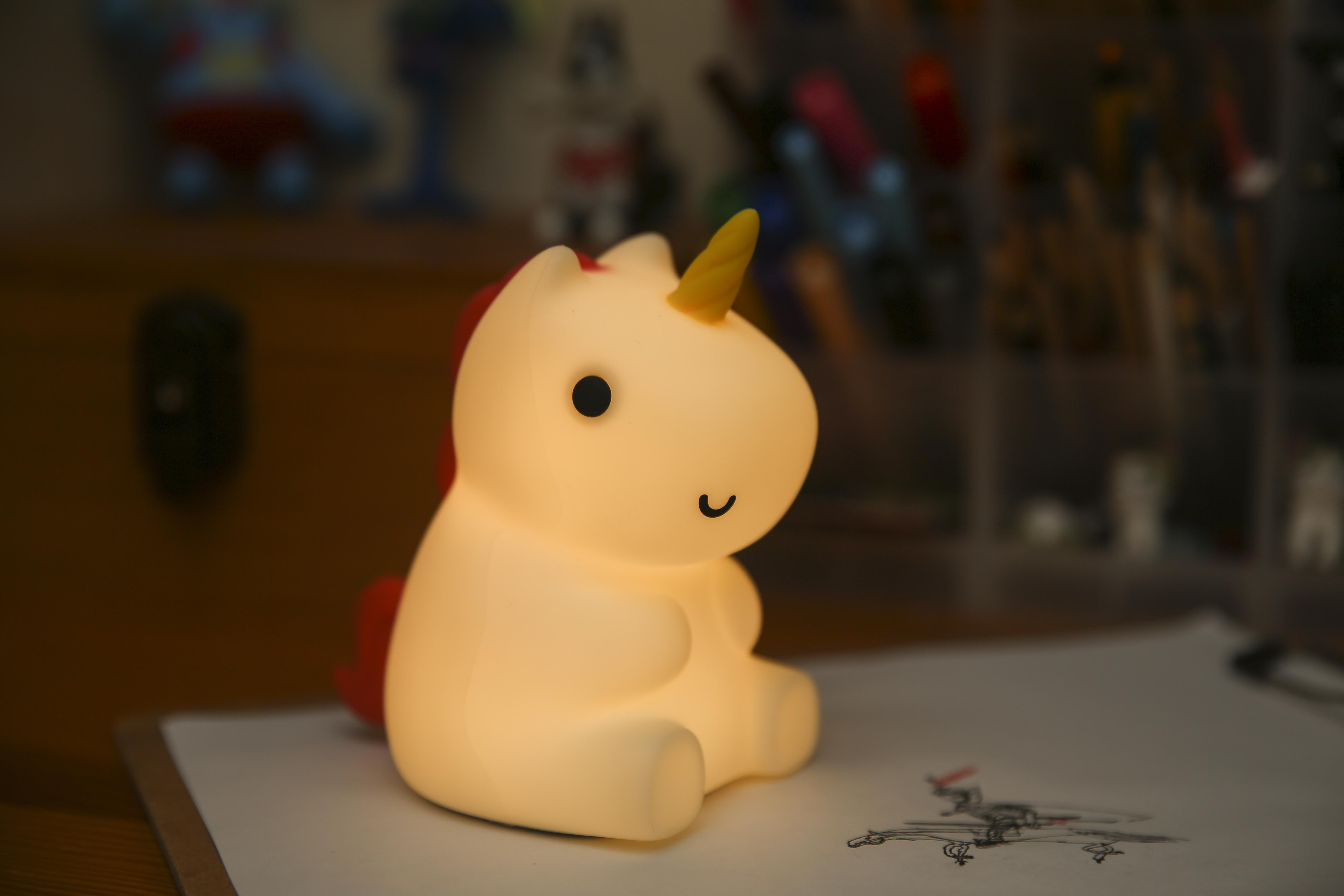 Leuchtendes Einhorn-Nachtlicht. Weißer Körper, gelbes Horn, rote Mähne und ein lächelndes Gesicht. Auf einem Schreibtisch.