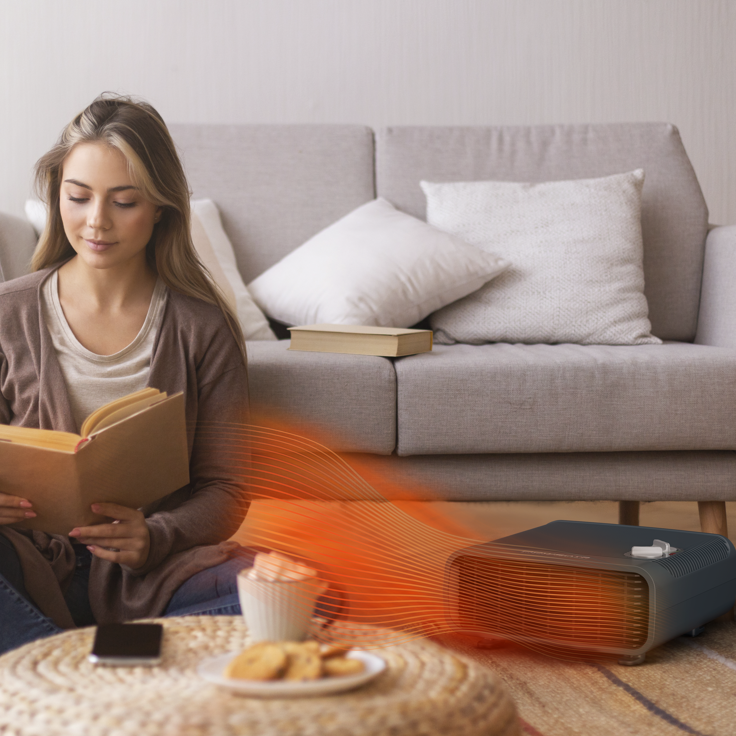 Mujer lee libro cerca de un calefactor. Galletas, teléfono y café en el suelo. Sofá y almohadas detrás.