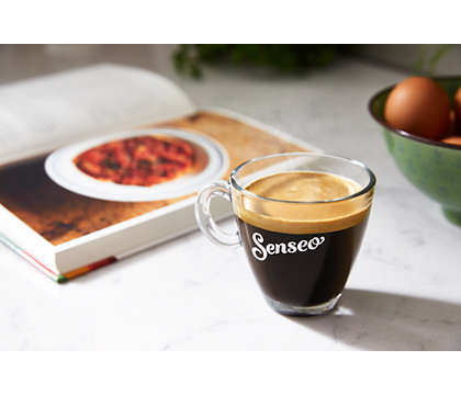 Koffie in een heldere glazen beker met een kookboek en eieren op de achtergrond.