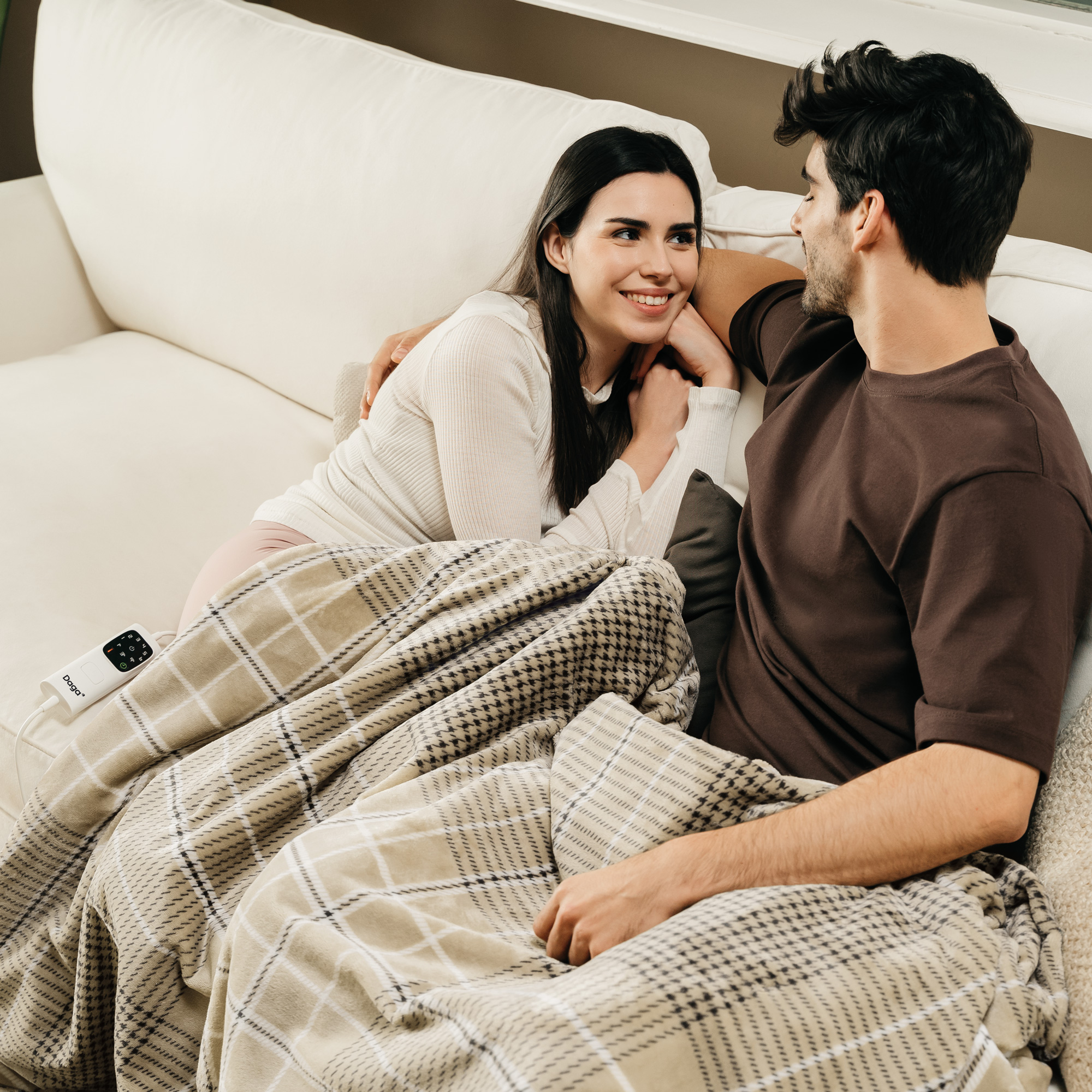 Pareja en el sofá, hombre y mujer abrazándose bajo una manta a cuadros. Decoración neutra.