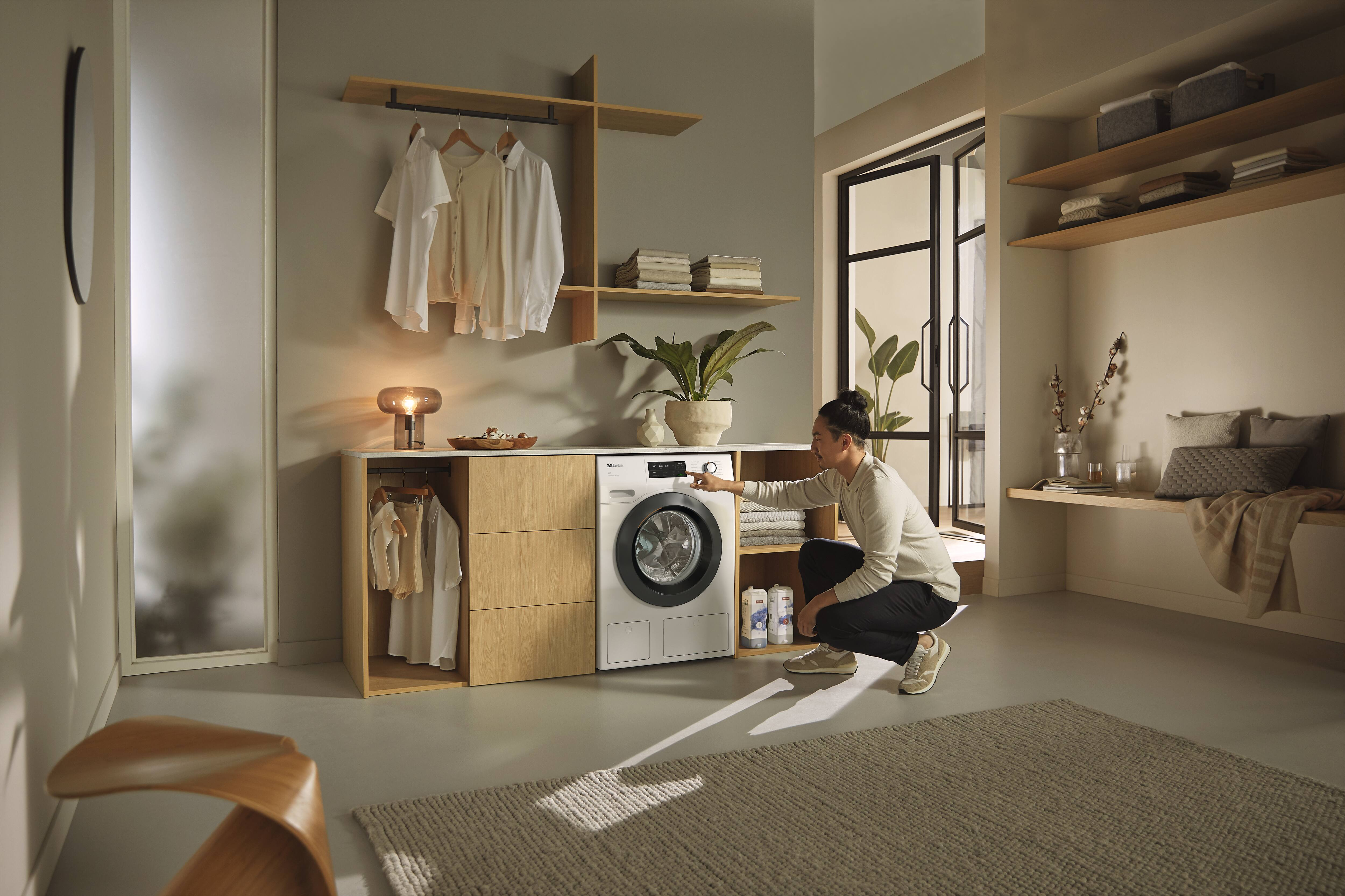 Une femme s'agenouille près d'une machine à laver, l'utilisant. Une étagère avec des vêtements est au-dessus d'elle.