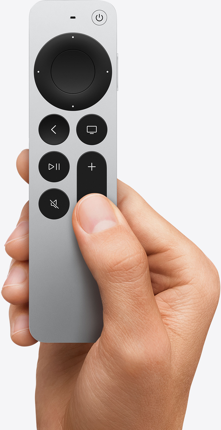 Kéz egy ezüst TV távirányítót tart, különböző gombokkal.