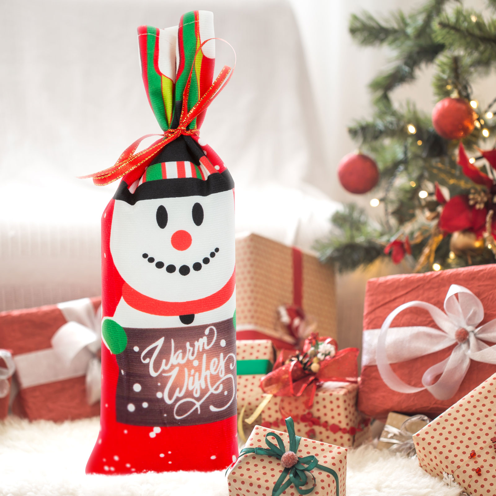 Piros hóember ajándéktáska 'Warm Wishes' felirattal, karácsonyi ajándékok a háttérben.