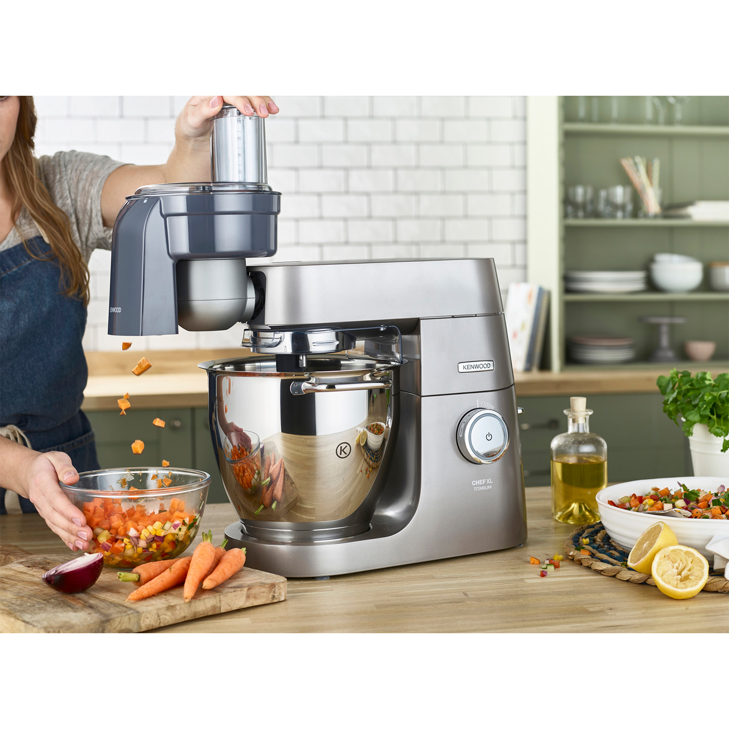 Una donna aggiunge ingredienti a un mixer da cucina argentato. Carote e altre verdure sono in vista.