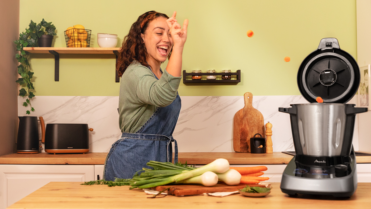 Mujer en la cocina riendo mientras lanza zanahorias a un procesador de alimentos.