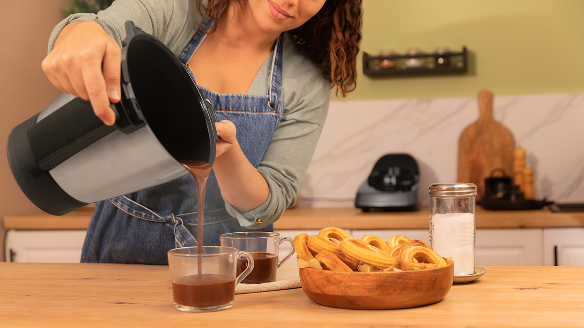 Mujer vertiendo chocolate caliente en dos tazas. Cuenco de madera con churros.