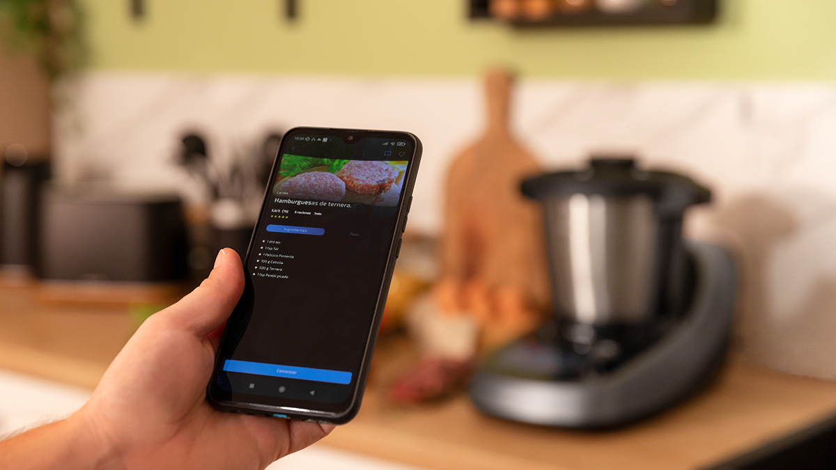 Mano sosteniendo un teléfono que muestra una receta. Un procesador de alimentos borroso en el fondo.