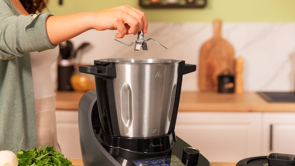 Una mujer coloca una cuchilla en un procesador de alimentos plateado. Entorno de cocina con hierbas verdes y tabla de madera.