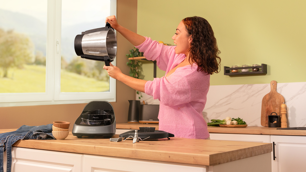 Mujer sosteniendo una batidora con cocina al fondo, sonriendo.