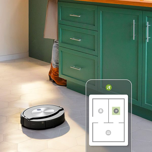 Ein Roboter-Staubsauger reinigt einen gefliesten Boden. Eine Person steht in der Nähe eines grünen Schranks.