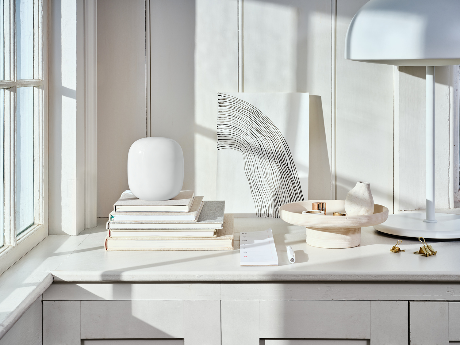 Een wit bureau met een witte lamp, boeken en een decoratieve vaas. Links een raam.