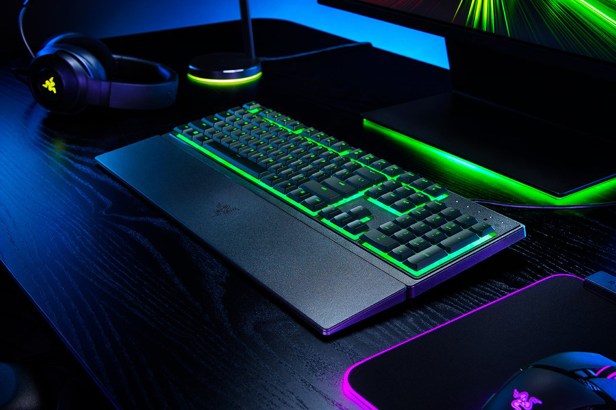 Fekete Razer billentyűzet zöld háttérvilágítással. A billentyűzet egy asztalon van, fekete egérrel és fejhallgatóval.