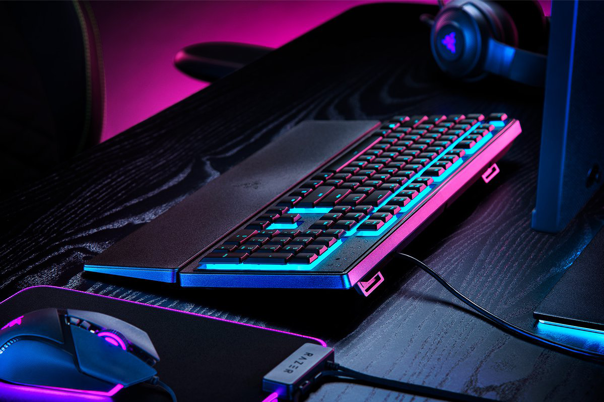 Egy gamer billentyűzet egy fekete asztalon. Rózsaszín és kék fények világítják meg.