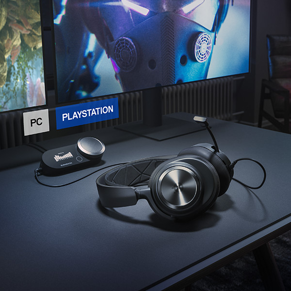A headset and control module on a desk. The background is a gaming setup with two screens.