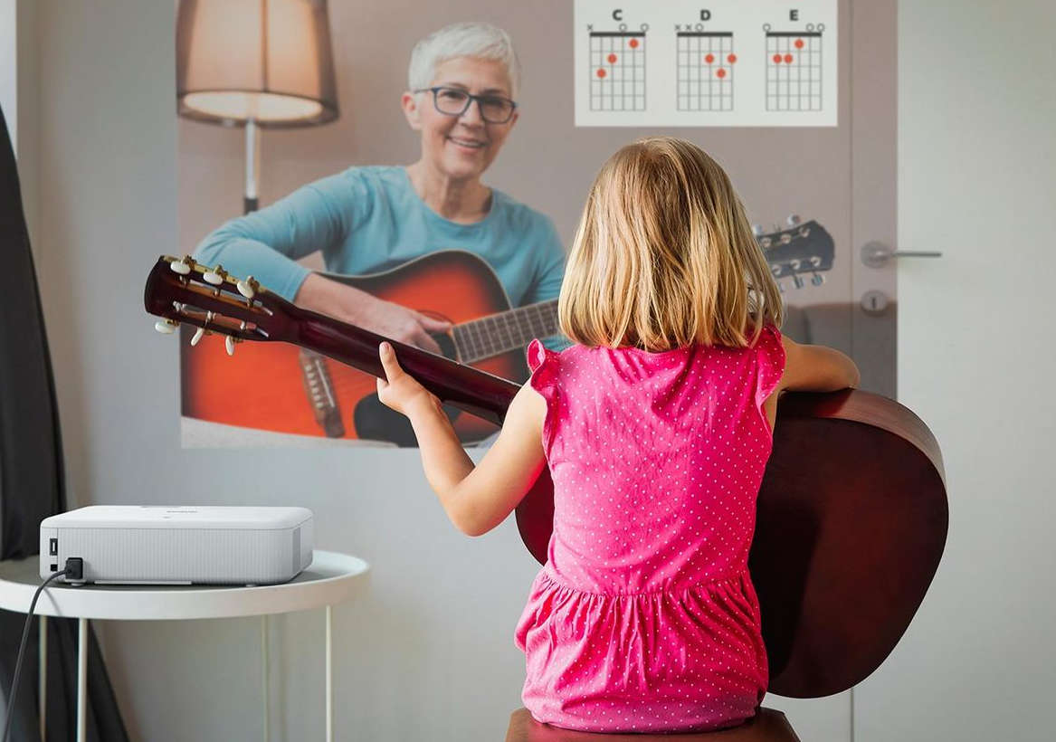 Una bambina impara a suonare la chitarra guardando una proiezione. Una donna anziana insegna dallo schermo.