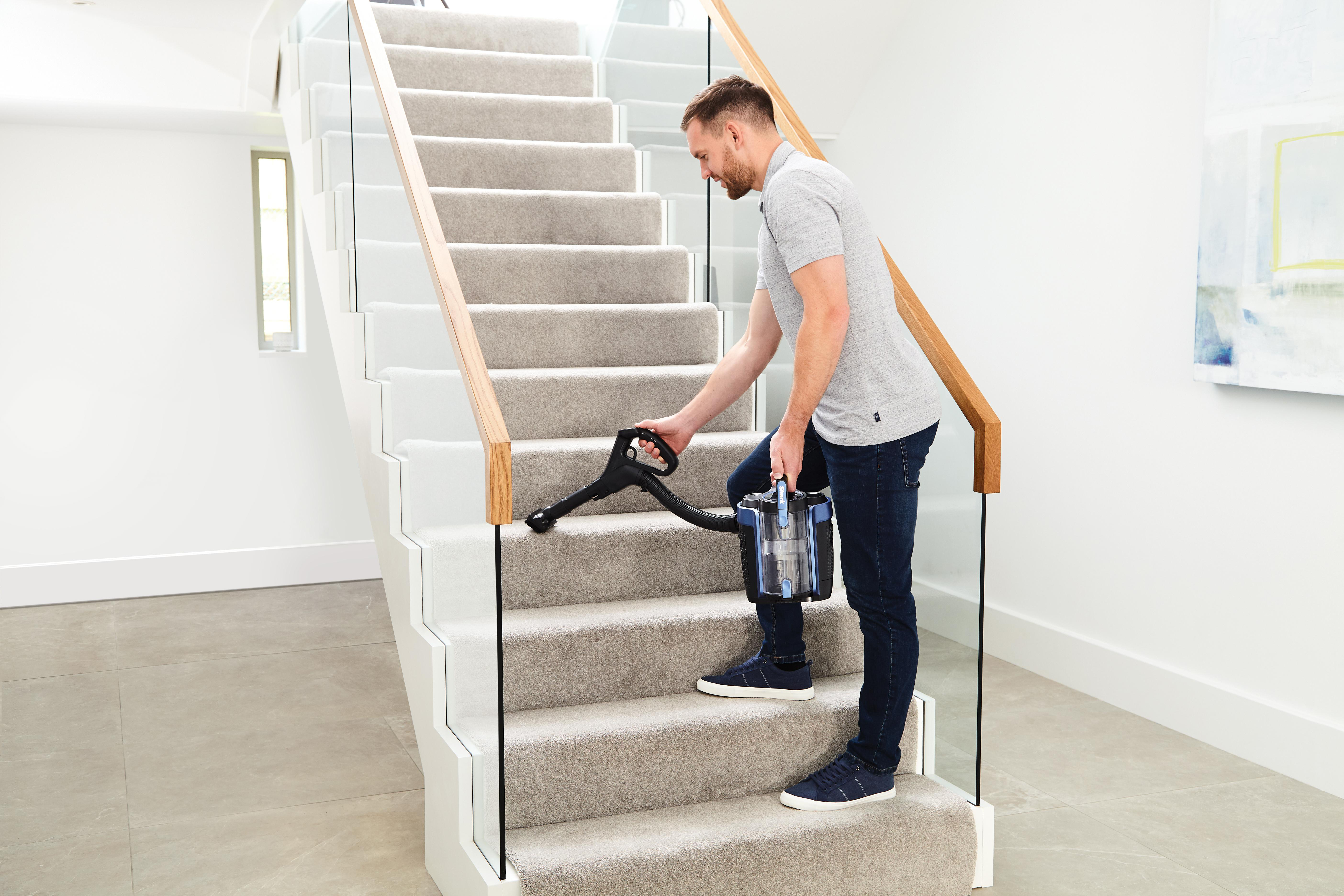 Mann saugt Treppen mit einem Handstaubsauger. Er trägt ein graues Hemd und Jeans.