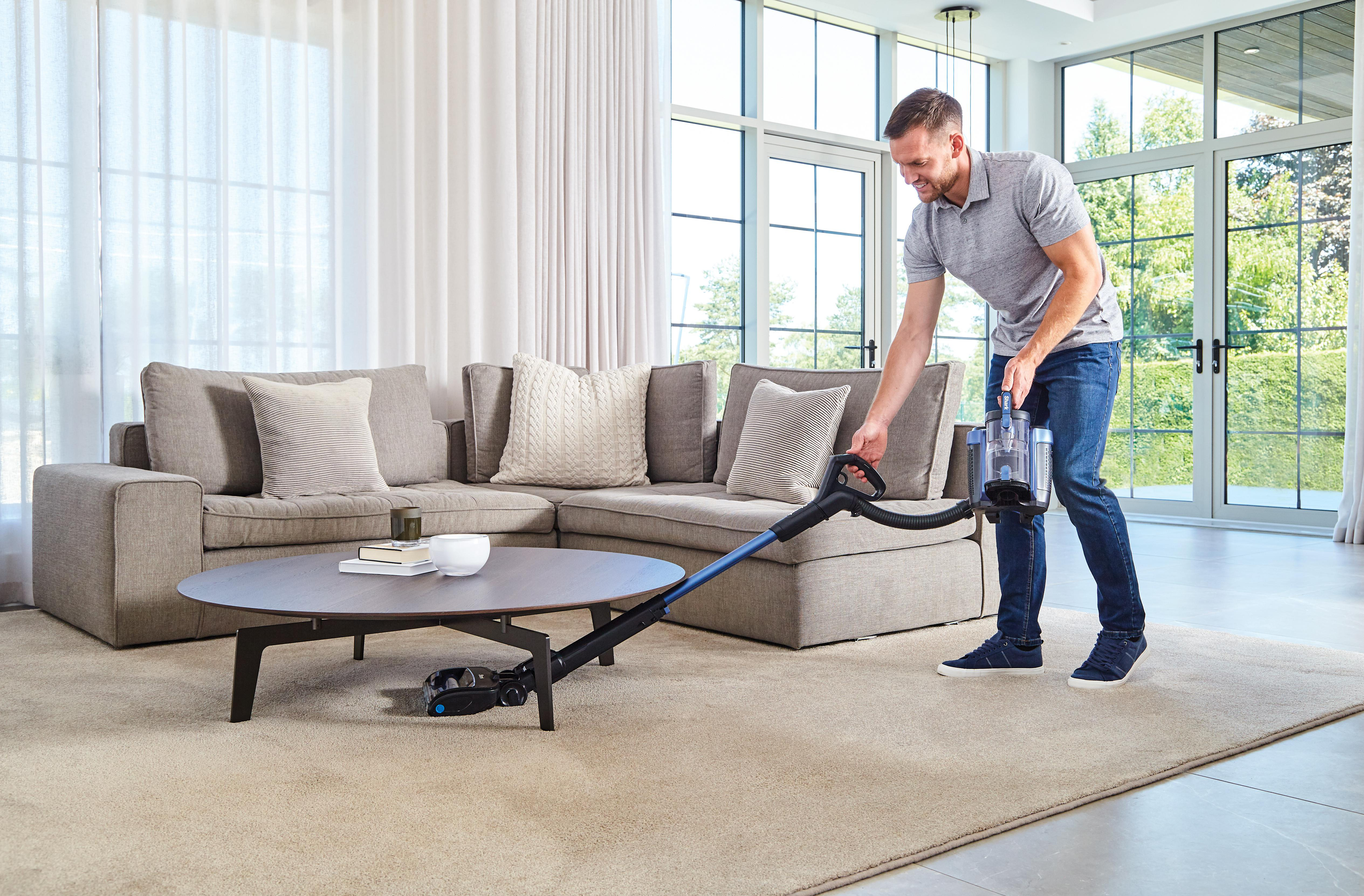 Mann saugt Wohnzimmer mit blauem Staubsauger. Sofa und Couchtisch sind im Hintergrund.