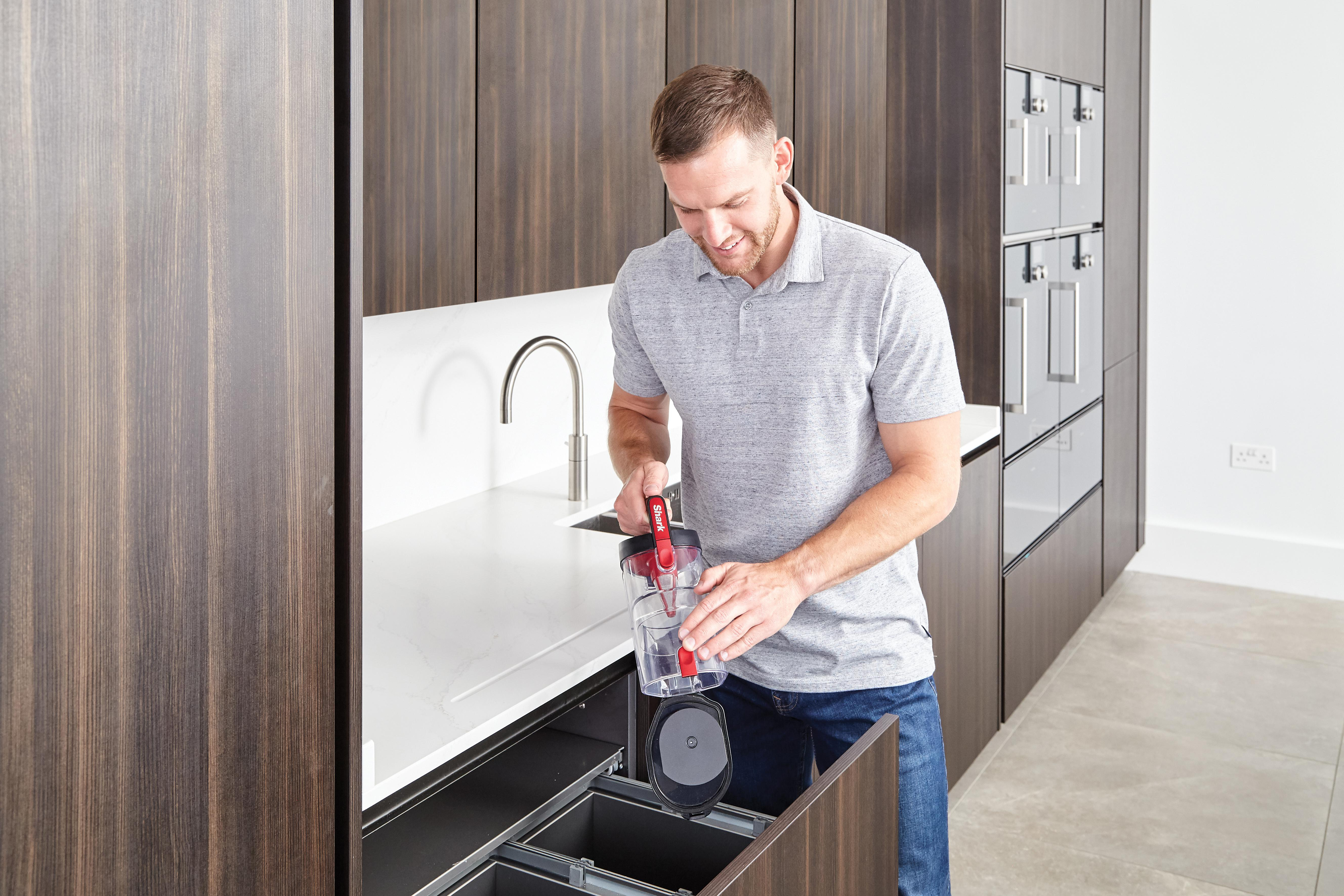 Mann in der Küche leert einen Staubsauger. Dunkle Holzschränke und weiße Arbeitsplatte im Hintergrund.