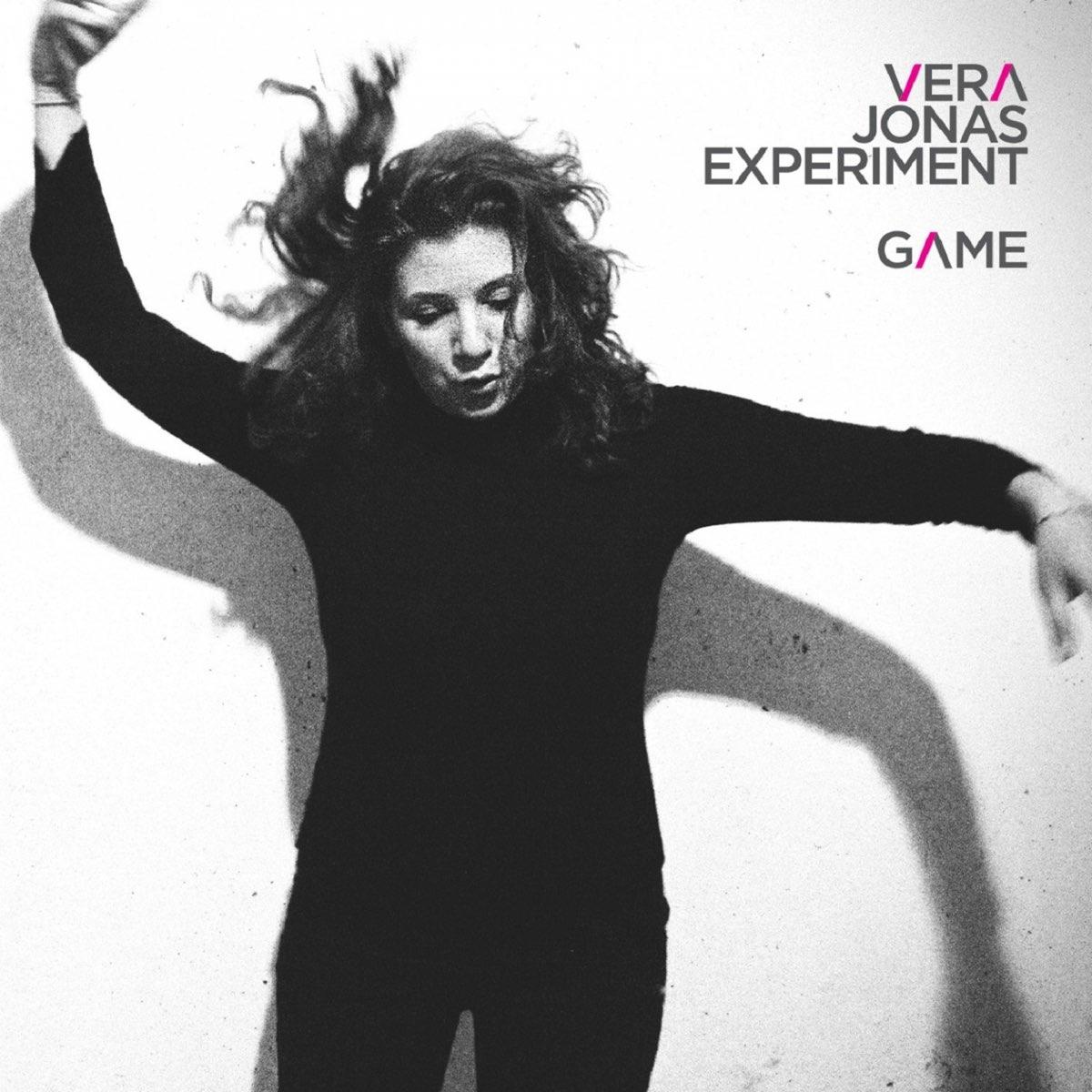 Fekete ruhás nő, karok felfelé, sötét haj. Szöveg: VERA JONAS EXPERIMENT GAME fehér falon.