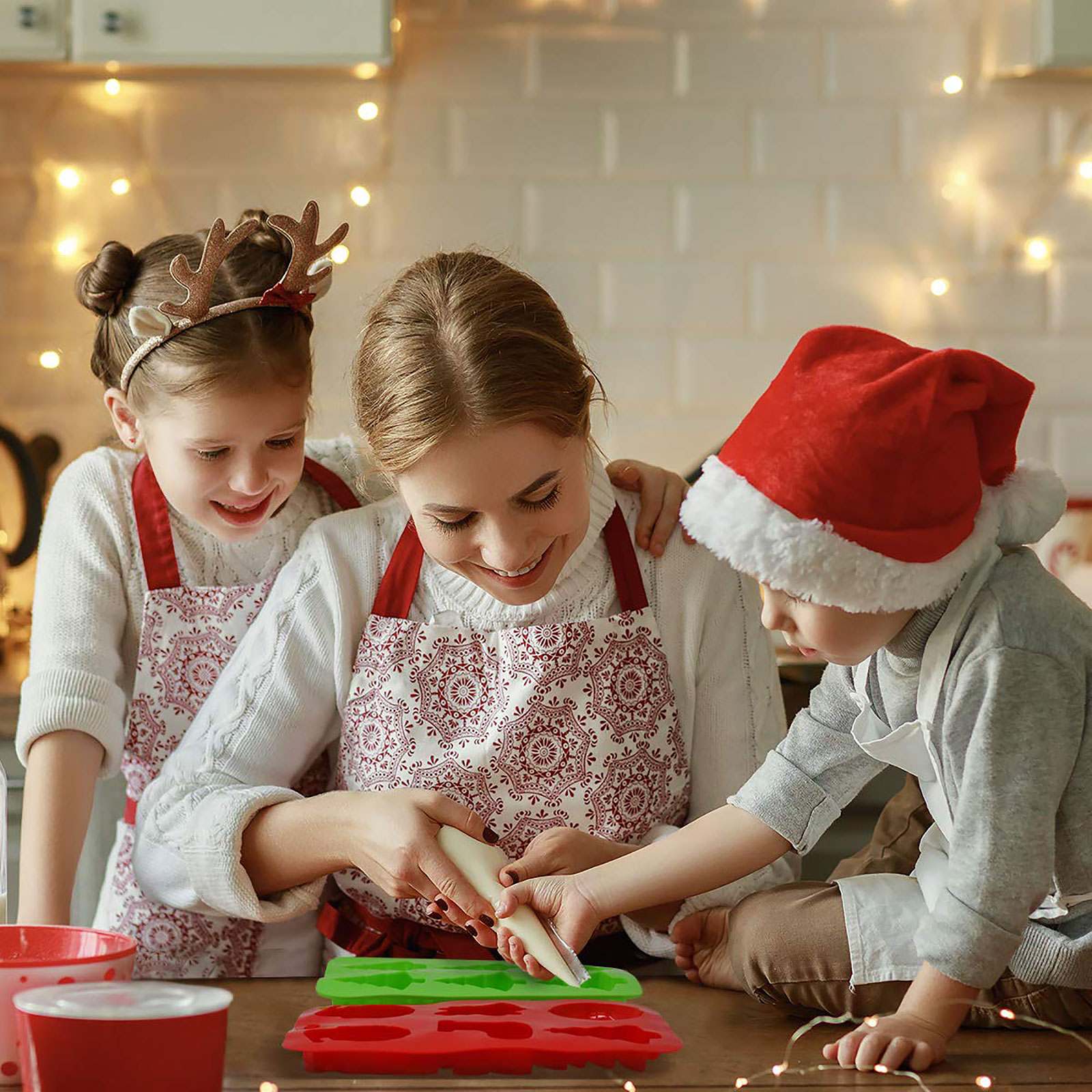 Család sütiket díszít: anya, két gyerek, piros és zöld süti formák, ünnepi környezet.